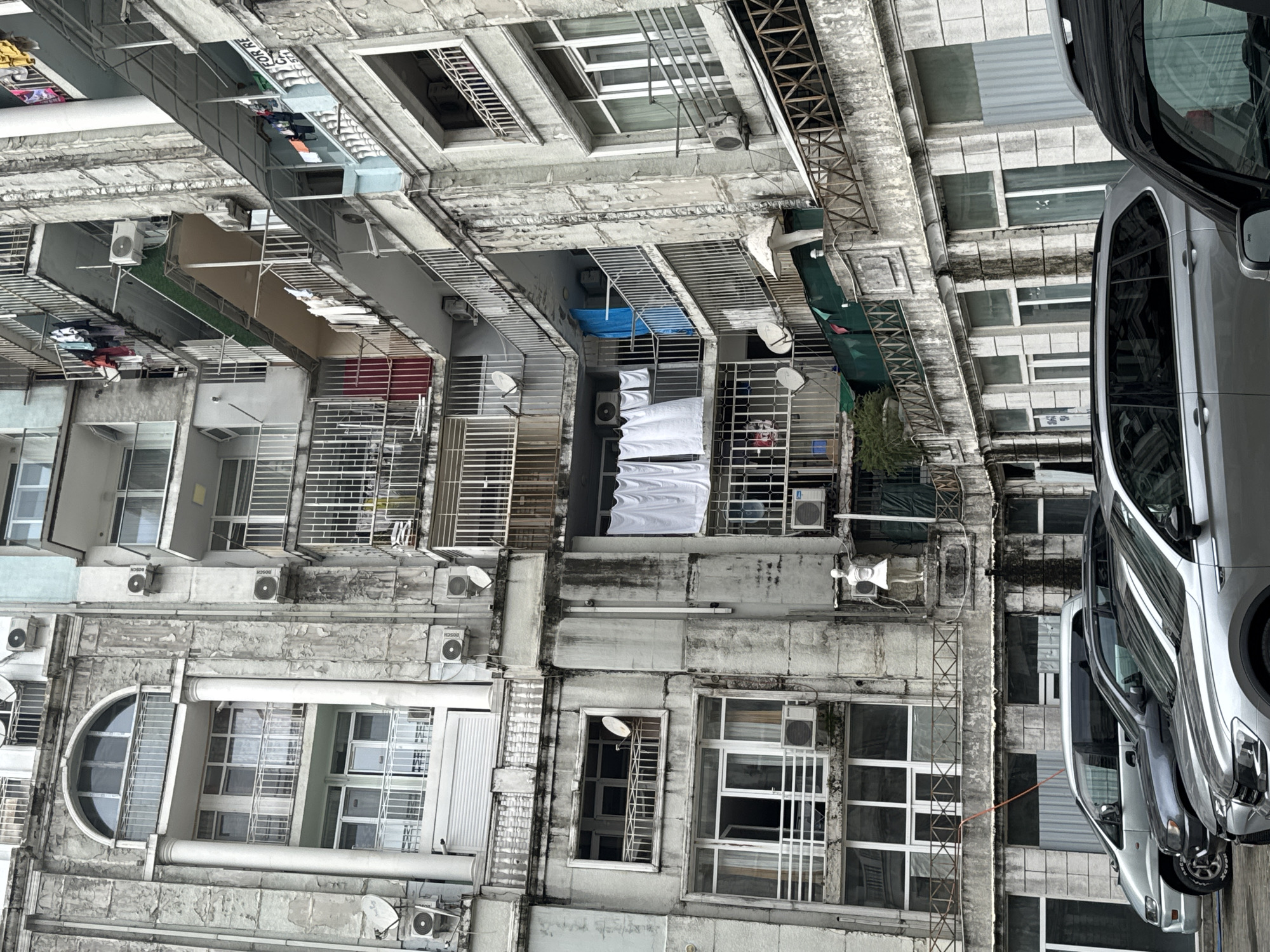 This is a street-level, exterior shot of a weathered, multi-story apartment building in Batumi, Georgia. The building exhibits signs of age and neglect, with peeling paint and stained concrete facades. Numerous balconies are visible, many adorned with laundry hanging out to dry, indicating residential use. Air conditioning units are mounted on many exterior walls, and satellite dishes are also present.
In the foreground, several parked cars are visible, including a silver SUV on the left, a darker SUV in the middle, and the hood and windshield of a black SUV on the right. The building's architecture includes classical elements such as columns and arched windows on the left side. A white statue of a female figure stands on one of the lower balconies. The overall impression is one of a dense urban environment with older residential structures.