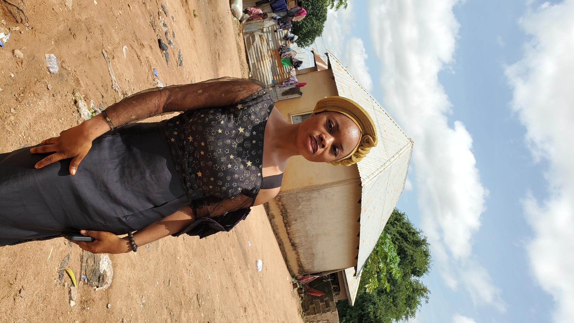The image shows a young woman posing in front of a building. She is wearing a gold headscarf and a black dress with gold star patterns. The building appears to be a residential structure with a corrugated metal roof. In the background, there is a clothesline with laundry hanging, and further in the distance, trees and a blue sky with clouds are visible. The ground is dusty and appears to be unpaved. This scene is consistent with a rural or semi-urban environment, potentially in a town like Jalingo, Nigeria, given the architectural style and general appearance of the surroundings.