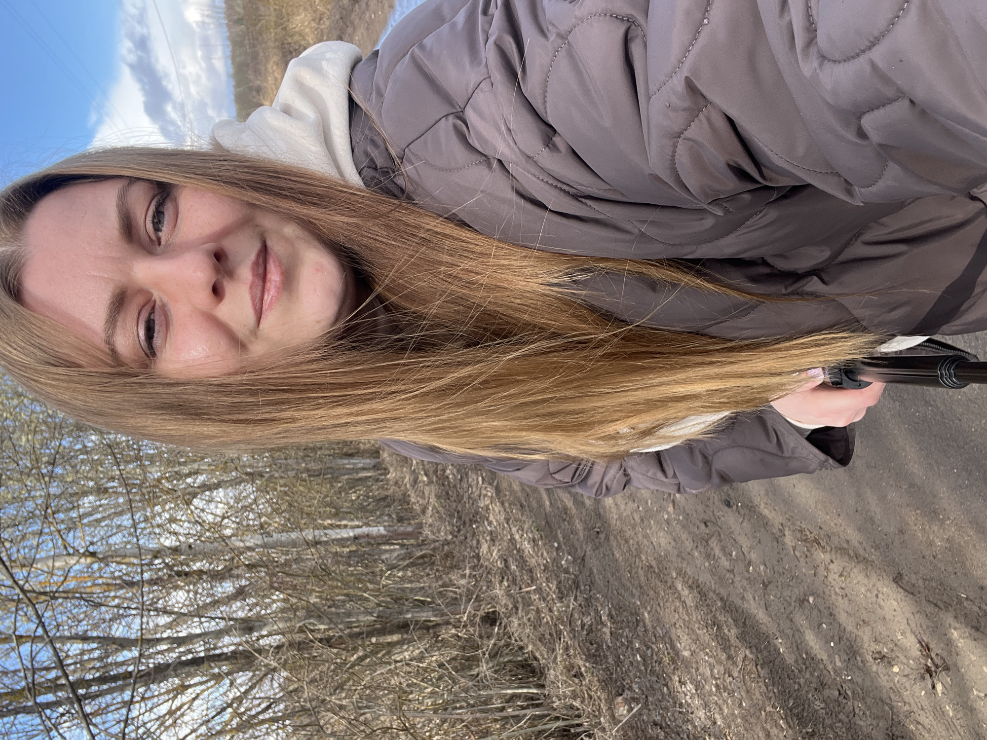 A woman with long blonde hair is taking a selfie outdoors in Opalevo, Russia. She's wearing a puffy grey jacket over a white hooded sweatshirt. The background consists of a dirt path with bare trees on one side and open sky with clouds on the other. The ground shows signs of dried leaves and twigs. She is holding a black selfie stick.