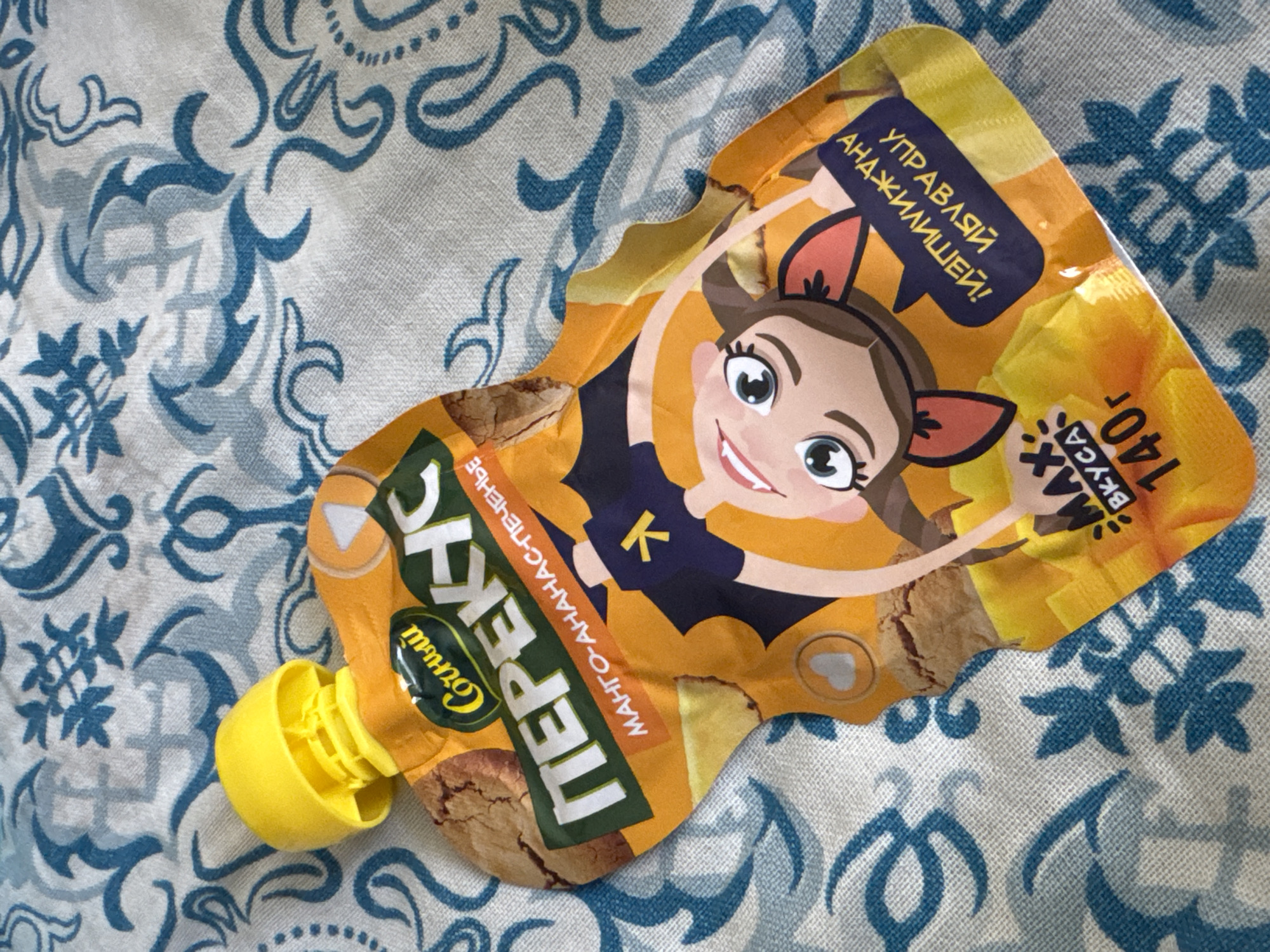 A pouch of baby food sits on a textured fabric surface with a blue and white pattern. The pouch is orange and yellow, with a cartoon illustration of a smiling girl wearing a dark blue outfit with a yellow "k" symbol on it. The pouch has text in Russian, including "ПЕРЕКУС" (snack) and "МАНГО-АНАНАС-ПЕЧЕНЬЕ" (mango-pineapple-cookie). Below the illustration, there is text in a blue speech bubble that reads "УПРАВЛЯЙ АНАЖИМЕИ!" which appears to be a playful or nonsensical phrase. Another section of text indicates "МАХ ВКУСА 140г" (Max taste 140g). A yellow cap is visible at the top of the pouch. The fabric surface appears to be a tablecloth or blanket. The image is a close-up shot of the baby food pouch, with the background slightly out of focus.