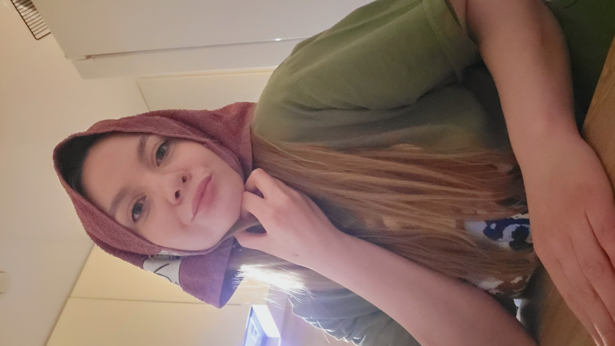 A woman with fair skin, blonde hair, and a pink towel wrapped around her head is visible in the image. She's wearing an olive green t-shirt and is resting her head on her hand, which is placed on a wooden table. The background is a light-colored wall with a glimpse of a white cabinet. The location appears to be indoors, possibly in a domestic setting. The city of Chaikovsky, Russia, is not discernible from the image.