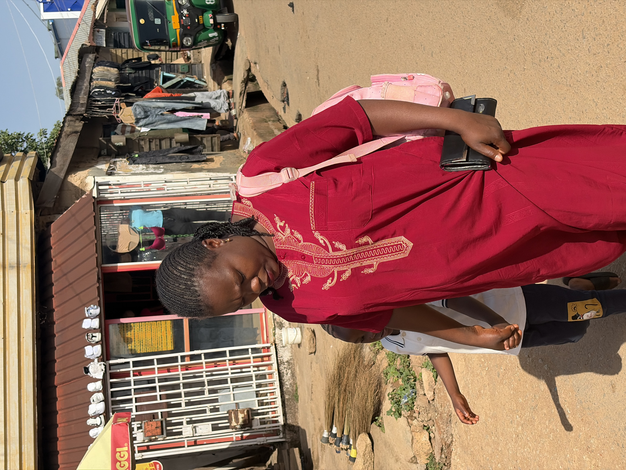 The image shows a woman in a red dress standing on a street in Jos, Nigeria. She has braided hair and is wearing a pink backpack. She is holding a black wallet in her hand. Next to her is a child in a white shirt and dark pants. Brooms are leaning against the side of a building. A small green vehicle is in the background. The buildings nearby have a reddish tin roof.