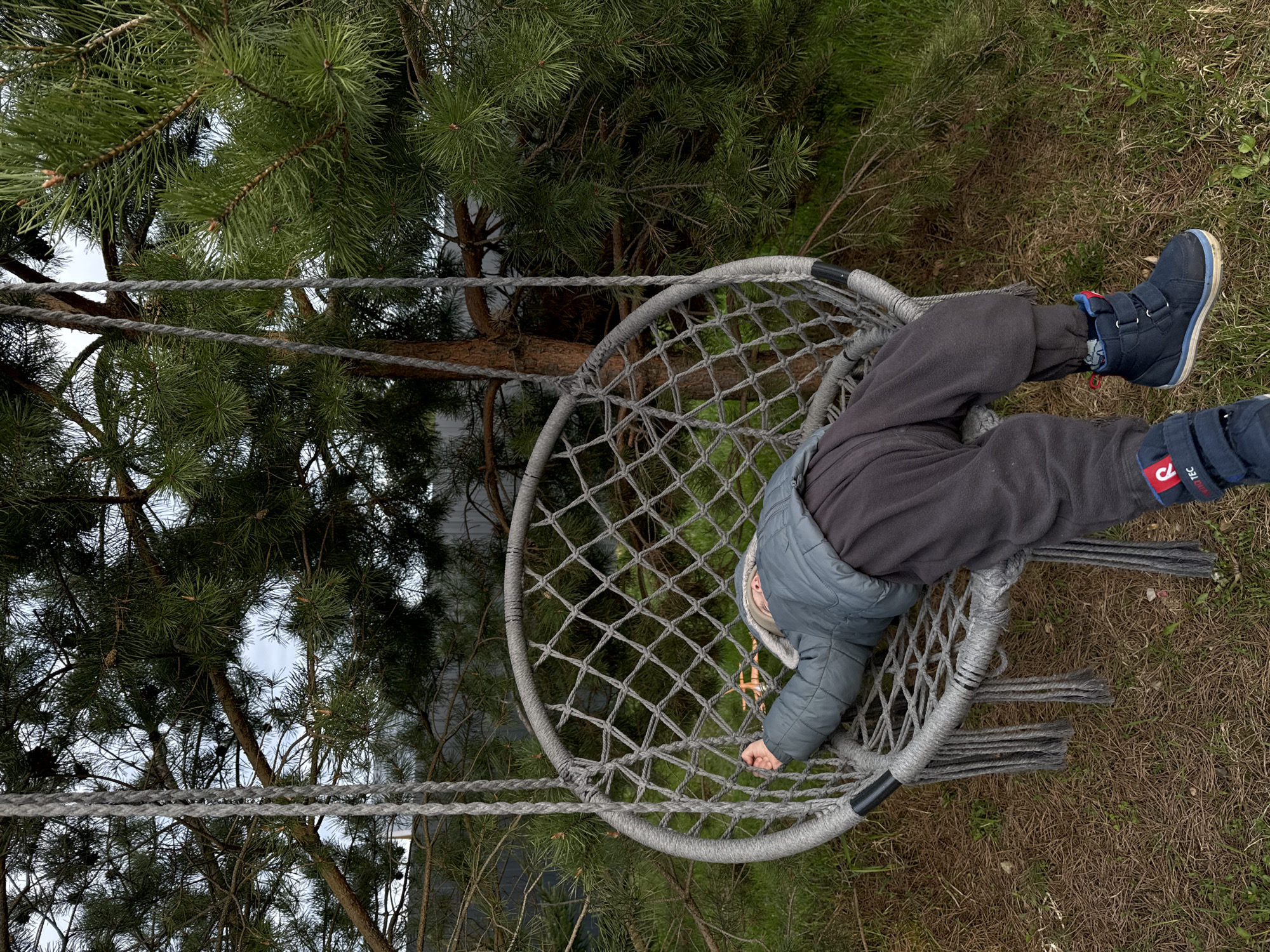 A young child is seen relaxing in a grey rope net swing, suspended outdoors amongst pine trees. The image, originally oriented on its side, shows the child lying down in the circular net, with their head to the left and feet extending to the right. They are dressed in dark grey sweatpants, blue winter boots, and a grey padded jacket. One of the child's arms reaches down towards the mesh of the swing.
The swing itself is crafted from thick grey rope, forming a sturdy net and outer ring. The background is dominated by lush green pine trees with visible brown trunks and needles, suggesting a natural, possibly forested, setting. Glimpses of a bright, likely overcast, sky can be seen through the tree branches. The ground beneath the swing is covered in dry grass and scattered pine needles.
Visible text on a red tag on the child's right boot or pant leg reads "TEC," possibly part of a brand name like Reima Tec. The scene appears to be taking place during the daytime, under soft, diffused light, indicating an overcast day. The location context provided is Litvinovo, Russia, aligning with the natural outdoor environment depicted. The child's relaxed posture suggests comfort and calm engagement with their surroundings.