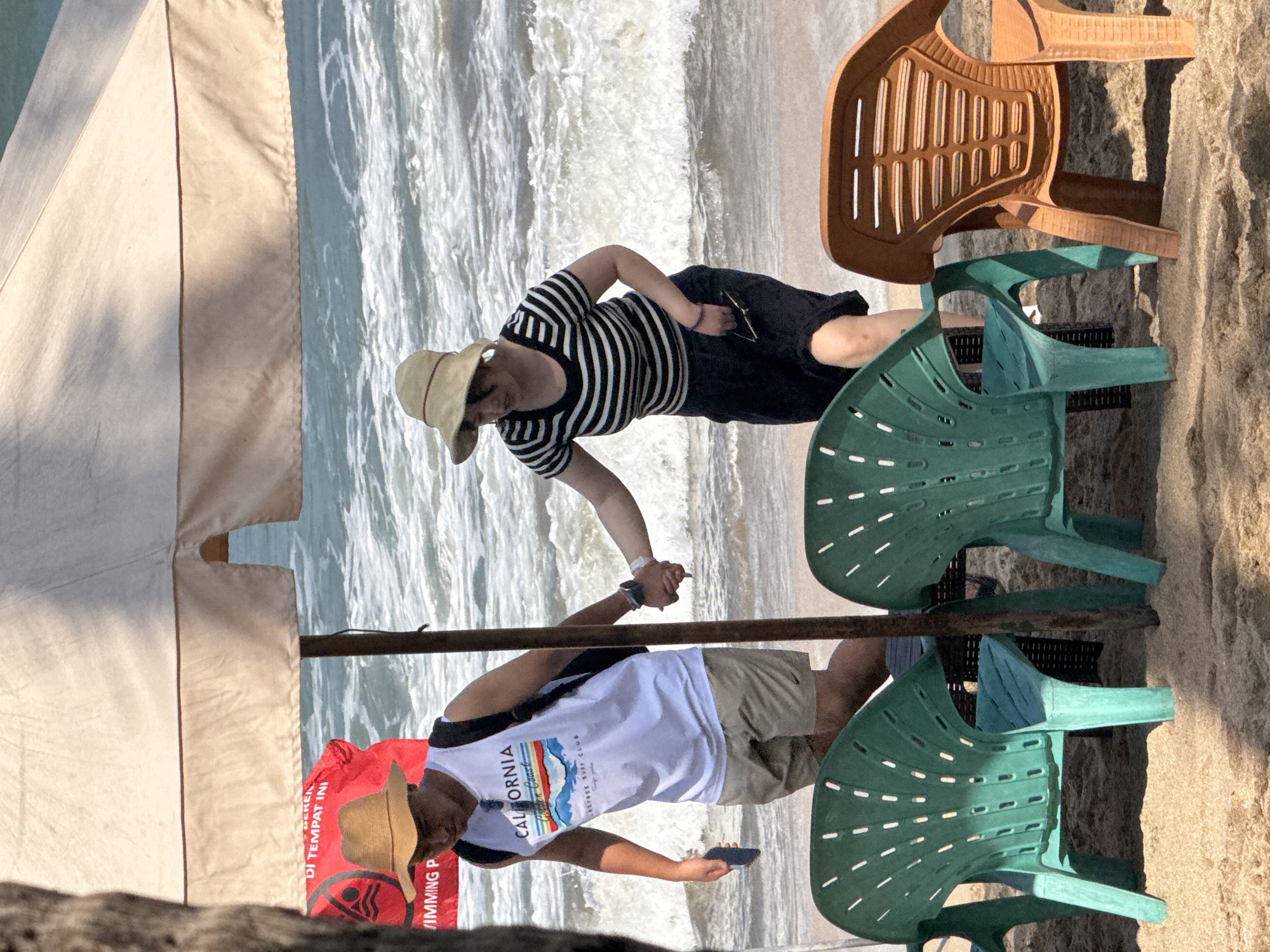 In Kuta, Indonesia, on a sandy beach adjacent to the ocean, a man and a woman are walking hand-in-hand under a large, light-colored beach umbrella. The man, wearing a straw hat, a white tank top with "California Golden Coast" printed on it, and khaki shorts, carries a backpack and a dark object in his right hand. The woman, also wearing a straw hat, a black and white striped shirt, and a dark skirt, holds a pair of sunglasses in her left hand. Two green plastic chairs are positioned in the foreground, and an orange plastic chair is to the right. The ocean in the background features moderate waves. A red sign with Indonesian text is partially visible behind the man.