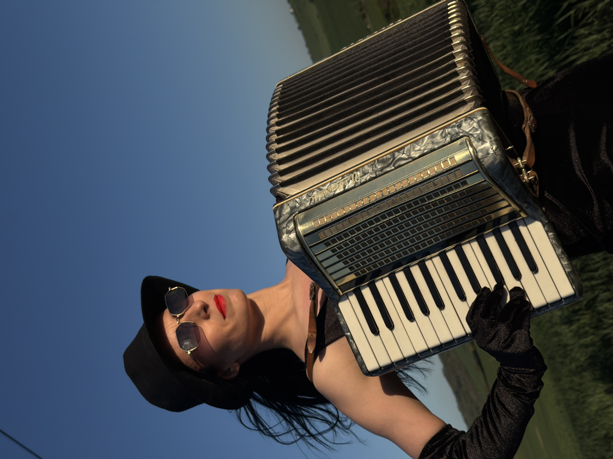 A woman in a black hat and sunglasses is holding an accordion, seemingly outdoors with a blue sky as the backdrop. Her red lipstick and dark outfit suggest a vintage or theatrical style. The scene appears to be in a field, with green grass visible. There's no information to determine whether the location is in Pleven, Bulgaria.
