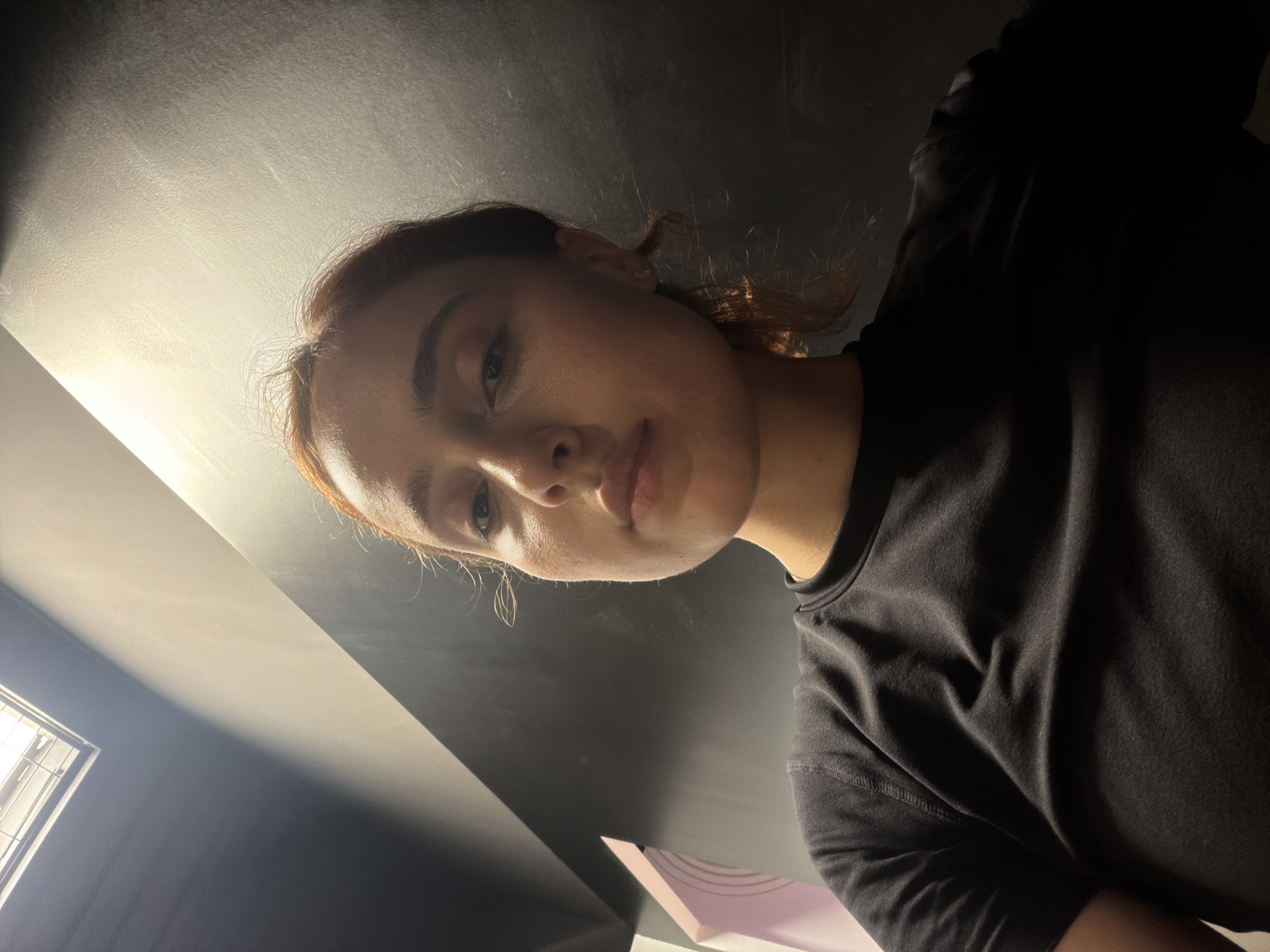 A young woman with reddish-brown hair is seen from a low angle, looking upwards towards the camera. She is wearing a black t-shirt and is positioned in an indoor environment. The background is dark, with a large, light-colored surface above and to the right of her head. A rectangular light fixture is visible in the upper left corner of the image. A portion of a pink and white object is visible on the left side of the frame. The overall lighting suggests a dramatic, low-light setting. The location is not specified to be Strasbourg, France.