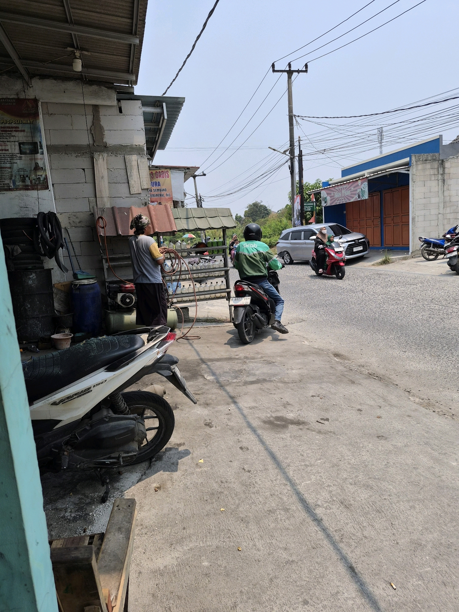In Wanajaya, Indonesia, on a sunny day, a street scene unfolds. A person wearing a camouflage bandana and a gray t-shirt is standing by a makeshift workshop, holding a red hose. Next to them, a blue barrel and a black compressor are visible. In the foreground, a white and black Honda motorcycle is parked. Further down the road, a silver minivan is parked in front of a building with blue siding and orange doors. A red scooter carrying two passengers is moving away from the camera. Another motorcycle, ridden by a person in a green jacket with a "Grab" logo, is also in motion. A blue motorcycle is parked on the right side of the street. The environment features concrete pavement, buildings, and overhead electrical wires.