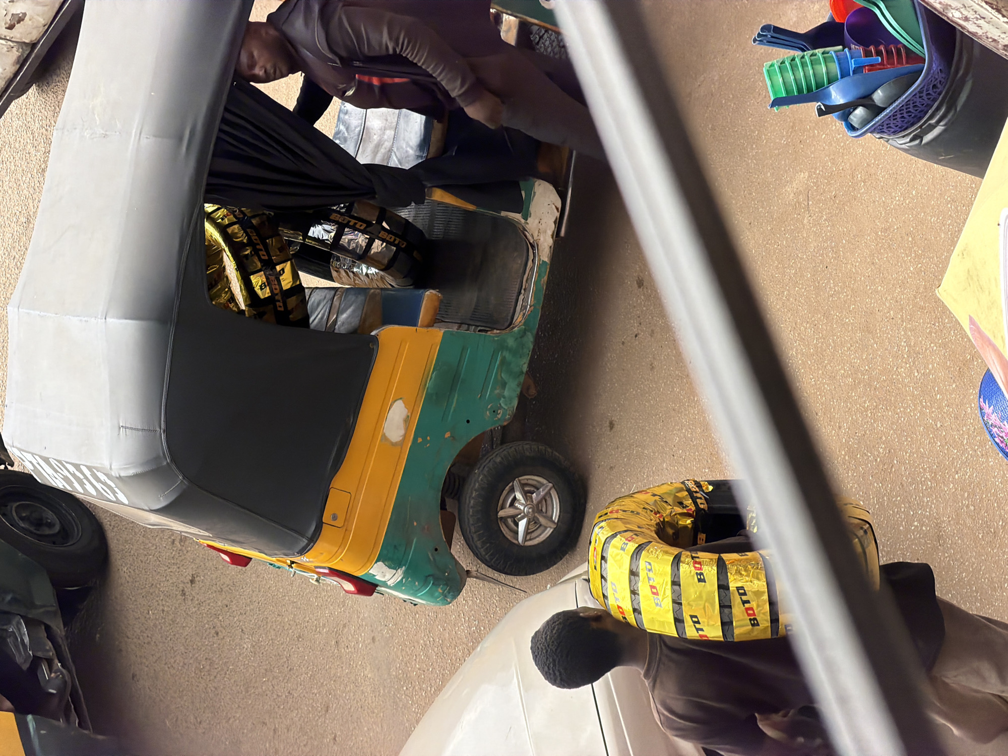In Bukuru, Nigeria, a three-wheeled auto-rickshaw, painted yellow and green, is parked on a paved surface. Inside the auto-rickshaw, a man wearing a dark brown jacket and pants is seated. In the foreground, a young person with dark skin and short hair is carrying a tire wrapped in bright yellow packaging with the word "BOTO" printed repeatedly in red. The packaging reflects light, making the tire appear golden. To the left of the auto-rickshaw, the side of a light-colored car is visible. In the lower right corner of the image, a black container holds a collection of colorful plastic kitchen utensils, including green cups, blue scoops, and a red object. The environment appears to be a street or a service area, with a gritty, sandy-colored ground.