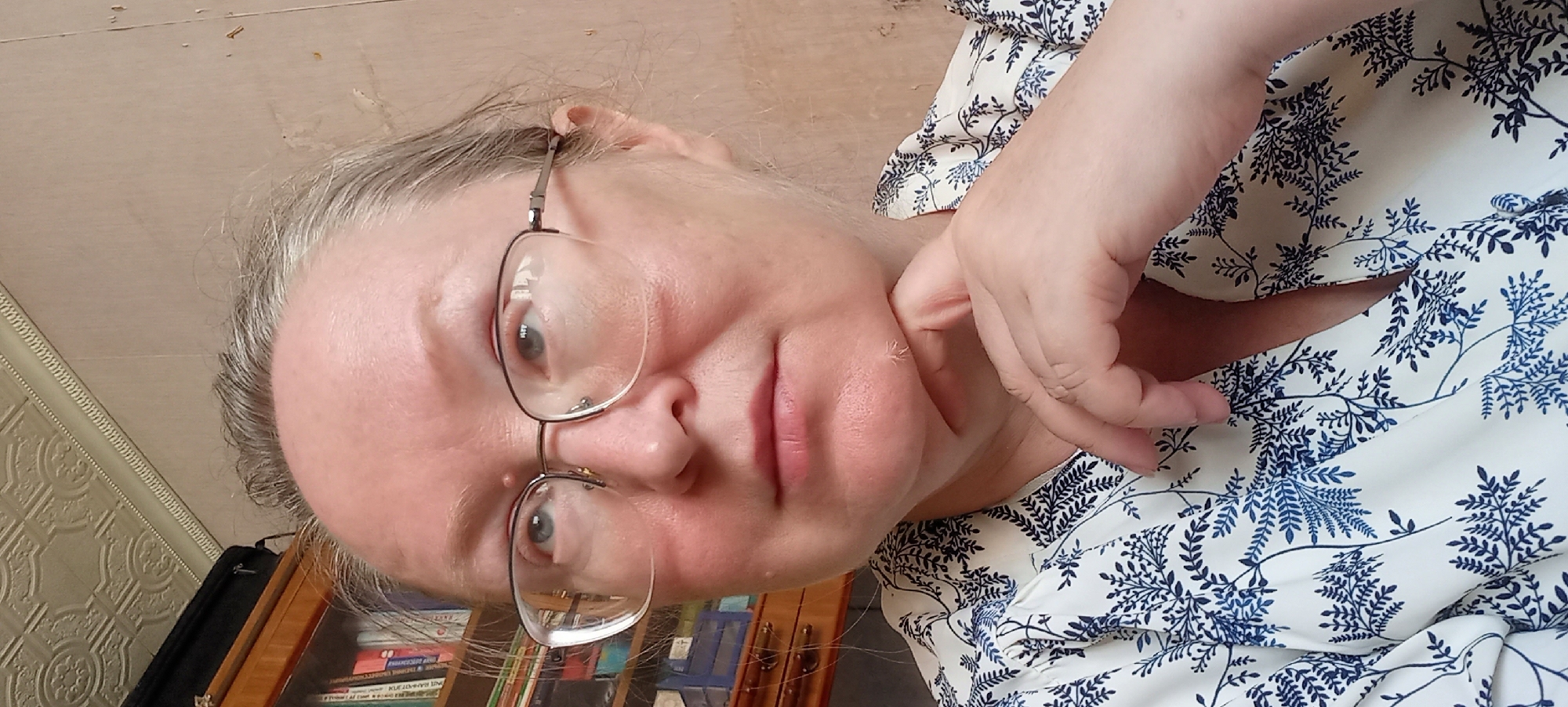 A woman with graying hair and glasses rests her chin on her hand. She is wearing a white blouse with a blue floral pattern. She is indoors, with a bookshelf visible in the background. The image is a close-up, centered on her face and upper torso. No specific location in Izhevsk, Russia, can be determined from the image.