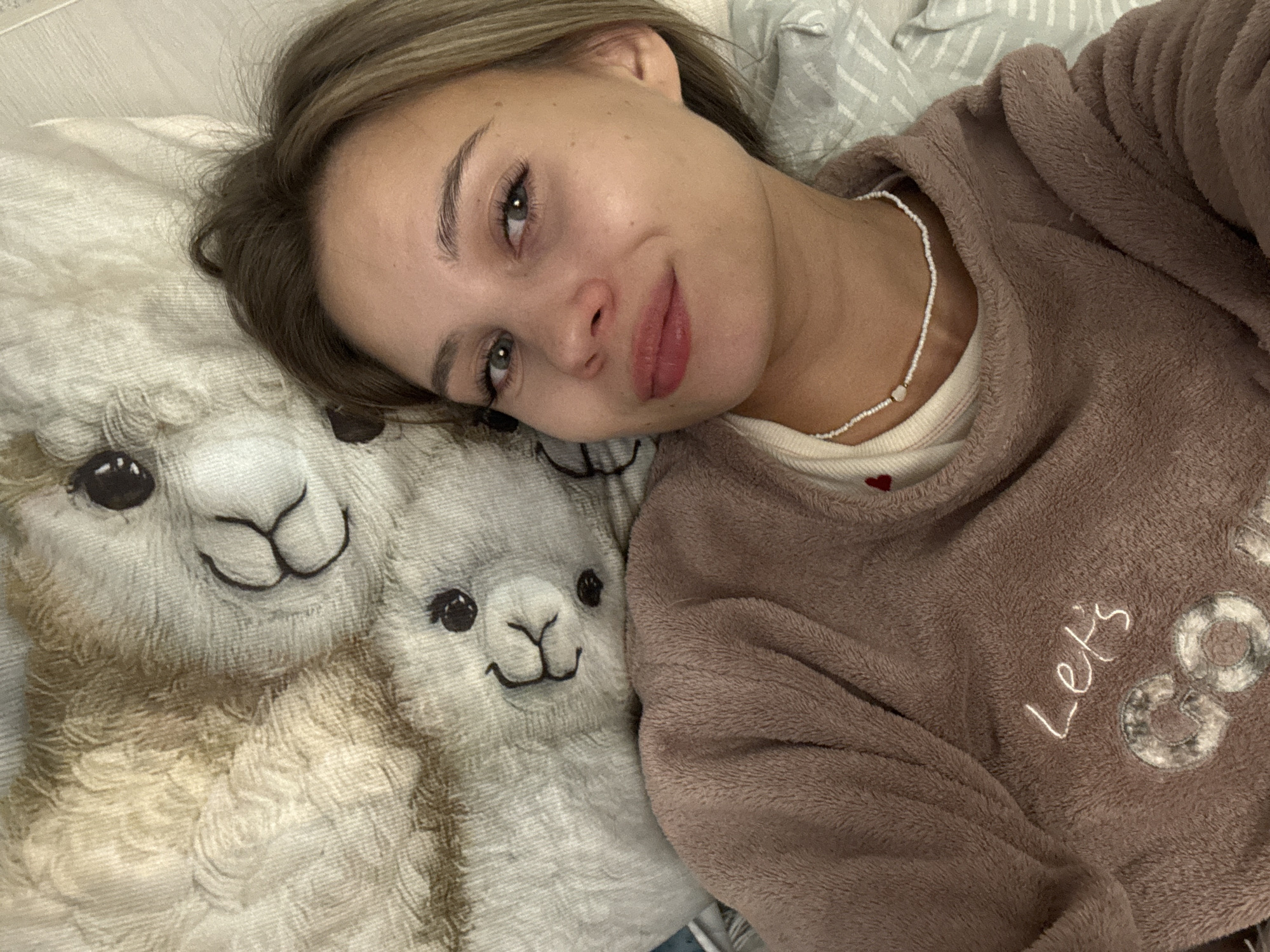 A young woman is lying on a pillow with two llama faces printed on it. She is wearing a brown fleece pajama top with the words "Let's Go" stitched on it. She has light brown hair and is wearing a beaded necklace. She appears to be indoors, likely in a bedroom, based on the bedding visible in the background. The image was likely taken in Chaikovsky, Russia.
