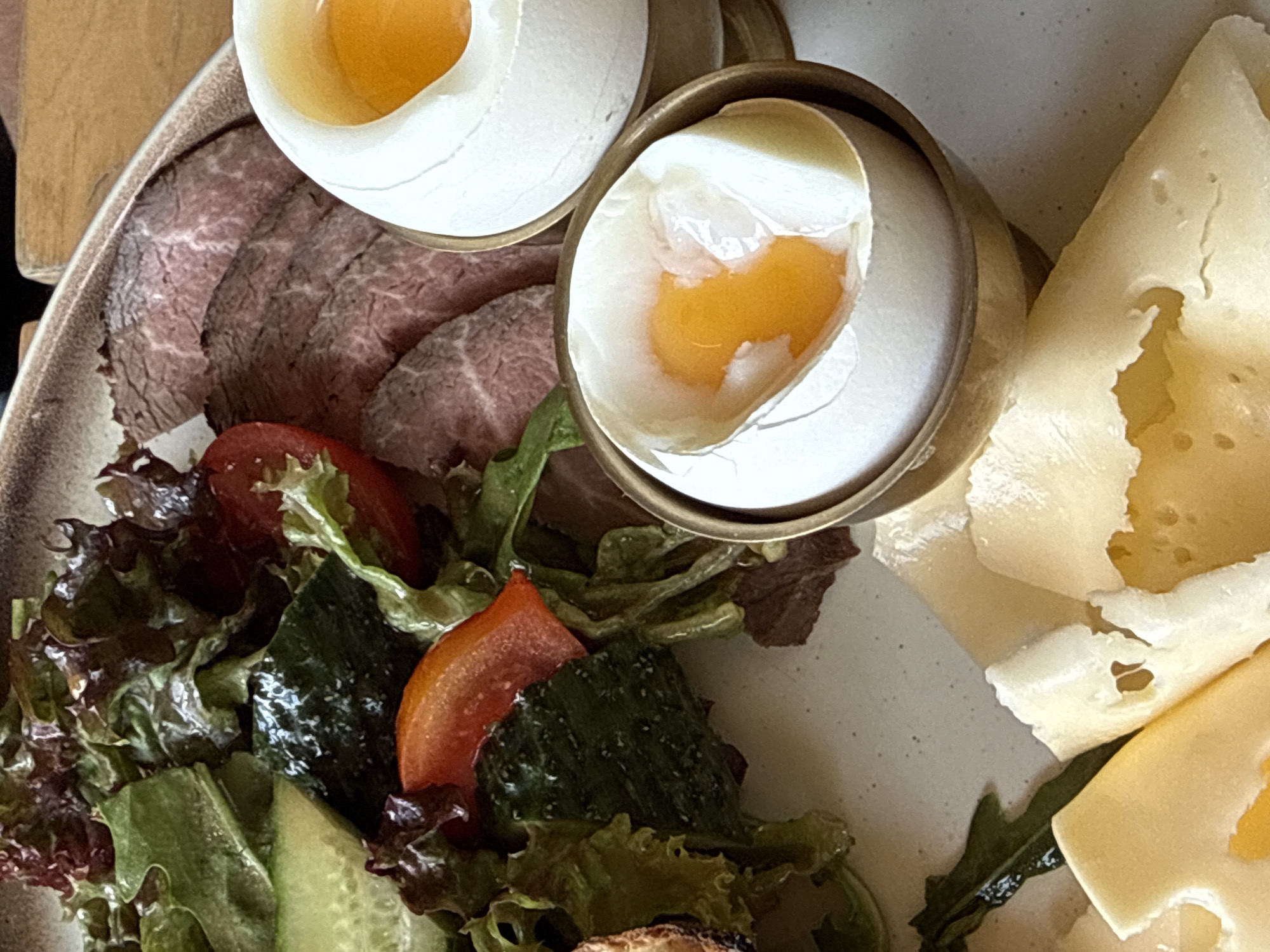 A close-up, eye-level, overhead shot displays a portion of a breakfast plate filled with a variety of food items. In the upper left quadrant, a vibrant salad is composed of red and green leafy lettuce, sliced cucumber, and chopped red bell pepper, with a few small tomato pieces. Adjacent to the salad, in the upper right quadrant, are two soft-boiled eggs, each served in a white egg cup with a gold rim. The eggs are open, revealing their runny, bright orange yolks. To the left of the eggs, thinly sliced roast beef is arranged on the plate. In the lower portion of the image, several slices of yellow Swiss cheese with characteristic holes are visible. The plate itself appears to be light gray with a speckled texture. The lighting is soft and diffused, highlighting the textures and colors of the food. The setting is a table, with hints of a wooden surface visible in the background.