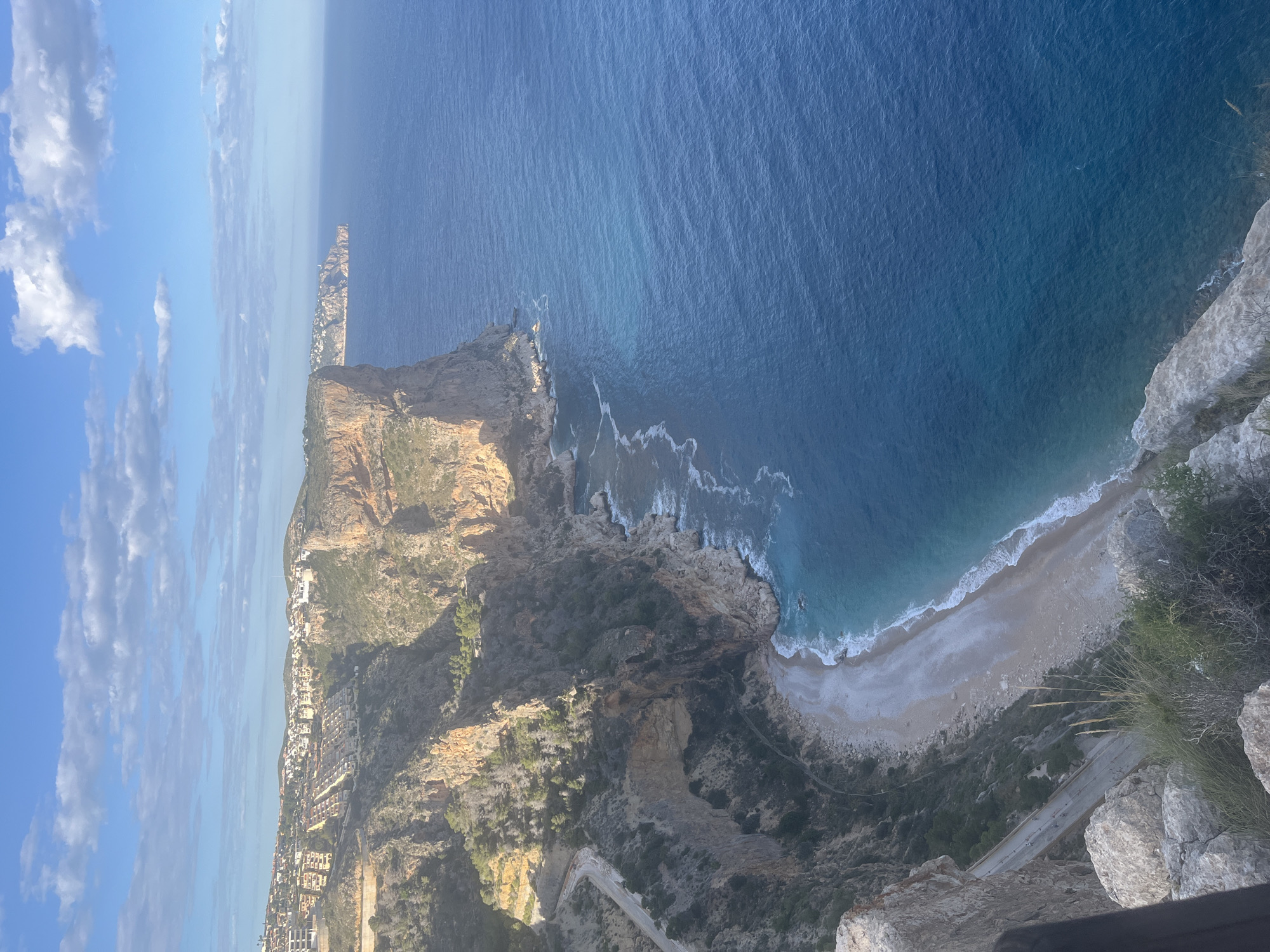 The image captures a high-angle, panoramic view of the coastline in el Poble Nou de Benitatxell/Benitachell, Spain.
Prominent features:
* **Coastline:** The rugged coastline includes a cliff edge, a sandy beach, and rocky formations meeting the water. Wave action is visible along the shoreline.
* **Water:** The sea displays varying shades of blue, indicating different depths or conditions.
* **Vegetation:** The landscape is covered in scrubby vegetation, which is most evident on the cliffs.
* **Built Environment:** Some buildings are nestled along the upper part of the cliffs towards the left side of the image.
* **Road:** A paved road is shown close to the cliff edge.
* **Sky:** The sky is blue with some wispy white clouds, suggesting sunny weather.
