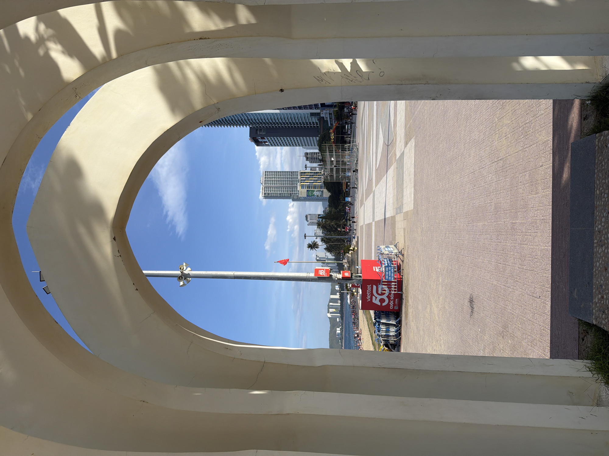 The image captures a view through a pale archway onto a sunlit urban plaza and beach in Nha Trang, Vietnam. In the foreground, the archway frames the scene with its curved, cream-colored structure, casting dappled shadows from unseen foliage onto its surface. The plaza itself is paved with patterned tiles in shades of beige and reddish-brown. To the left, a beach is visible with numerous small figures of people scattered across the sand, and further out, the sea stretches towards a distant coastline with low-rise buildings and hills.
Dominating the mid-ground is a tall, silver flagpole flying a red flag. Adjacent to the flagpole is a prominent red structure with white lettering and logos, including "Viettel" and "5G." Several rows of folded blue chairs are stacked behind this structure. Beyond the plaza, the city skyline rises with several modern high-rise buildings, their glass facades reflecting the bright blue sky. Palm trees line the street separating the plaza from the beach. The overall scene depicts a lively public space on a clear day, blending urban development with coastal leisure.