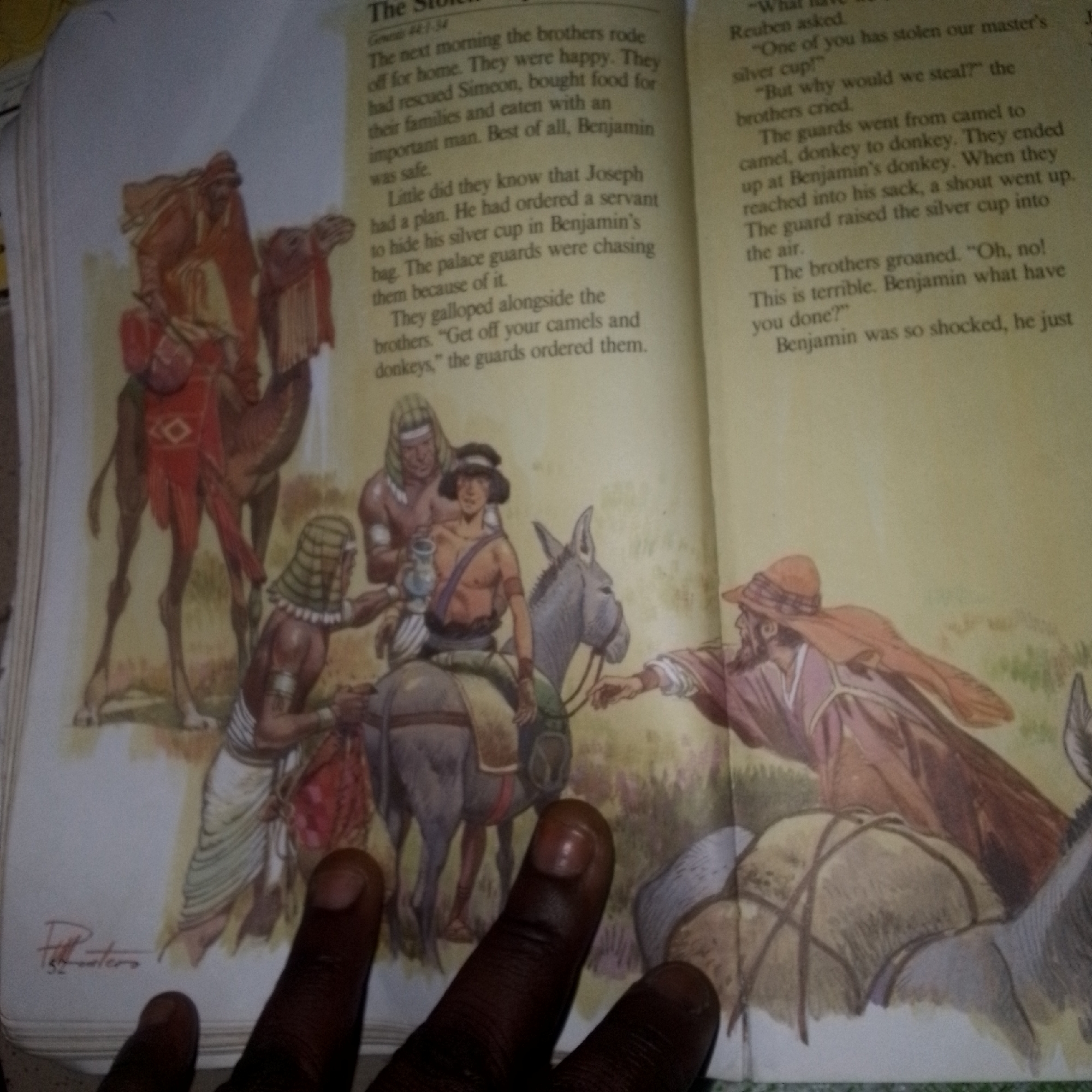 The image is a close-up, overhead shot of an open book displaying an illustration and text. The illustration depicts a scene with several figures in what appears to be ancient attire, some on camels and a donkey, with a man in a brown robe and hat prominent in the foreground. The text discusses a biblical narrative involving Joseph, Benjamin, and a stolen silver cup. The location is not specified in the image or the text. The book is laid open, and the right side of the frame is partially obscured by fingertips. The overall setting suggests a storybook or educational material.