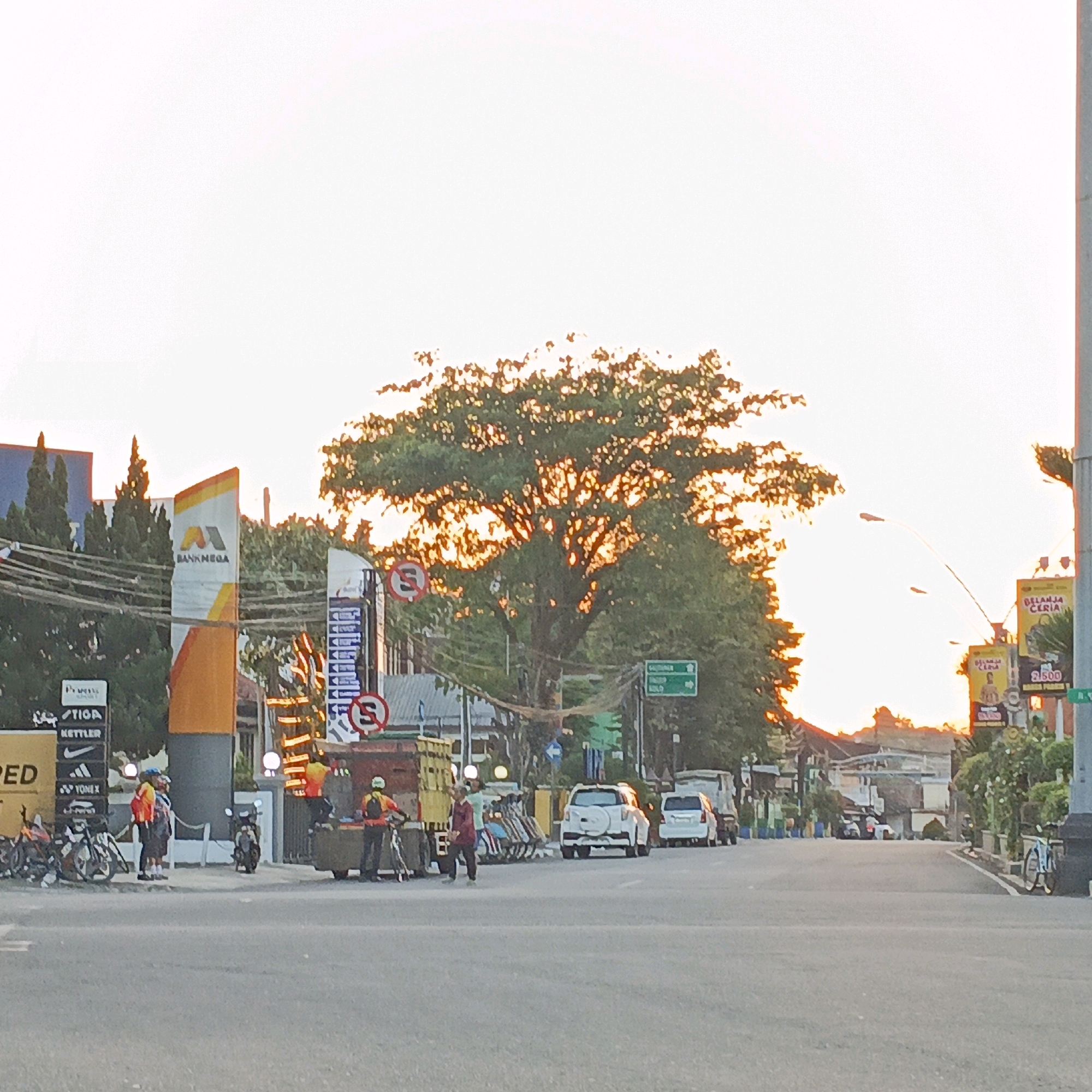 This image captures a street scene in Salatiga, Indonesia, during what appears to be late afternoon or early evening. The sun is setting, casting a warm glow through the leaves of a large tree dominating the center of the frame. The sky is bright and hazy, suggesting the sun is low on the horizon.
On the left side of the road, there's a Bank Mega building with its distinctive logo. Adjacent to it are several billboards and signs, including those for "TIGA" and "Kettler." Several motorcycles are parked near the sidewalk. Two individuals, possibly cyclists, are standing and talking near the parked motorcycles, one wearing a yellow and red jacket and the other a darker jacket.
Further down the road on the left, a large truck with a red cargo box is parked, and two people are near it. One person is wearing a yellow helmet and orange vest, holding a bicycle, while another individual in a maroon shirt is walking away from the truck.
On the right side of the street, several cars are visible, including a white SUV and another white vehicle further down. Traffic appears moderate, with vehicles moving in both directions. There are streetlights and various signs, including traffic signs with a red circle and a diagonal line, indicating prohibitions. Two large billboards on the right advertise "BELANJA CERIA" (Happy Shopping) with prices displayed. The road surface is asphalt, and the sidewalks are paved. Trees and smaller vegetation line parts of the street. The overall impression is of a busy urban street with the backdrop of a natural sunset.