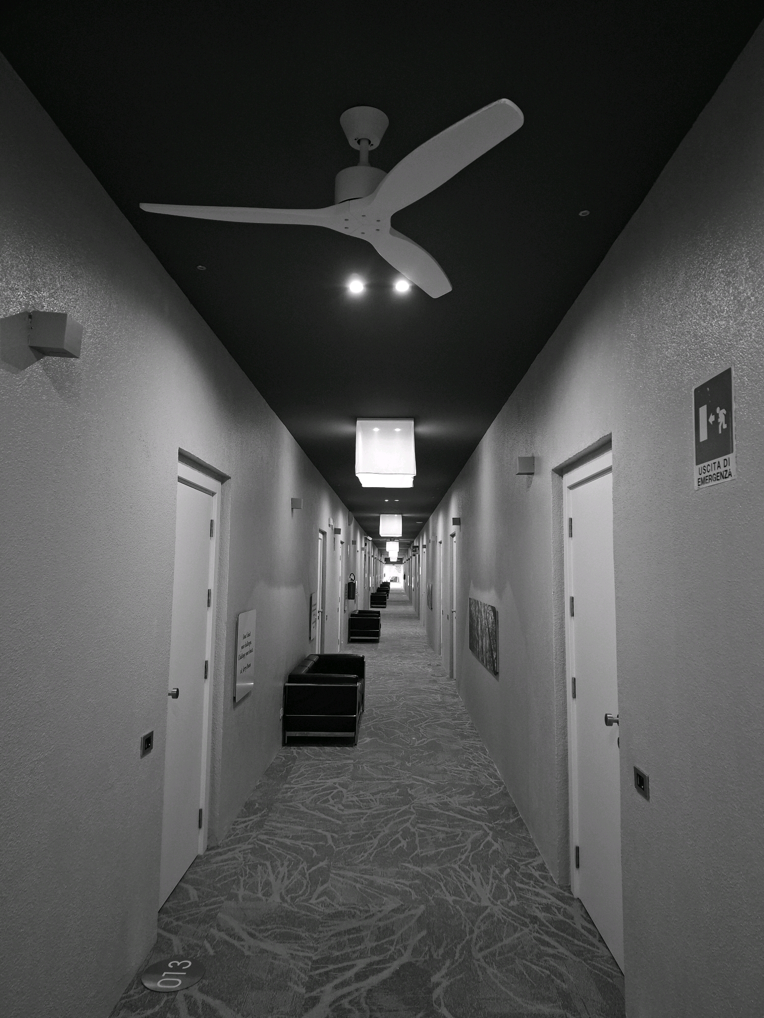 This is a grayscale, eye-level, medium shot photograph of a long, narrow hotel hallway. The hallway is dimly lit, with the exception of the ceiling lights and two spotlights directed at the ceiling fan. The walls are a textured, light gray, and the floor is covered with a patterned carpet. On the left wall, there are several white doors, each with a rectangular light fixture above it. On the right wall, there are also white doors, with similar light fixtures. Toward the end of the hallway, there is an emergency exit sign with the text "USCITA DI EMERGENZA." There are also several black lounge chairs placed along the hallway, and a painting on the right wall. The hallway appears to be empty of people. The overall impression is one of modern, minimalist design. The photograph does not provide enough visual information to determine the specific location of the city of Olgiate Olona, Italy.