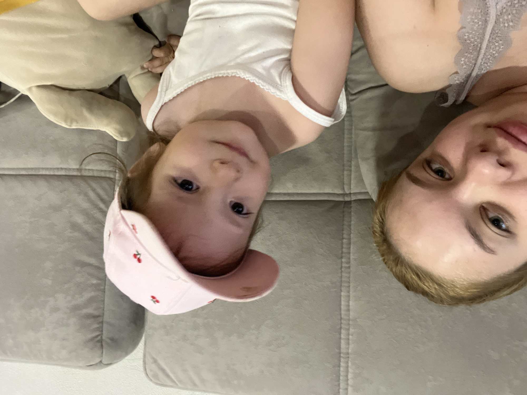 The media file is a selfie-style image taken indoors, likely in a home setting in Gorodets, Russia. Two people are visible, a young child and an adult, both lying down on what appears to be a sofa.
The child, in the foreground and on the left side of the image, is wearing a white tank top and a pink baseball cap adorned with embroidered cherries. The child is looking directly at the camera with a playful expression. To the right of the child is the adult, whose head is also angled towards the camera. The adult is wearing a light-colored, possibly gray, lace-trimmed top. Both individuals are positioned upside down relative to the viewer, suggesting the photo was taken from a low angle or that the camera was held in an unusual orientation.
The background consists of a textured gray sofa. There are no clear indications of the time of day or weather. The scene is intimate and casual, suggesting a relaxed moment between the child and the adult. No visible text is present in the image. The overall mood is one of warmth and familial connection.