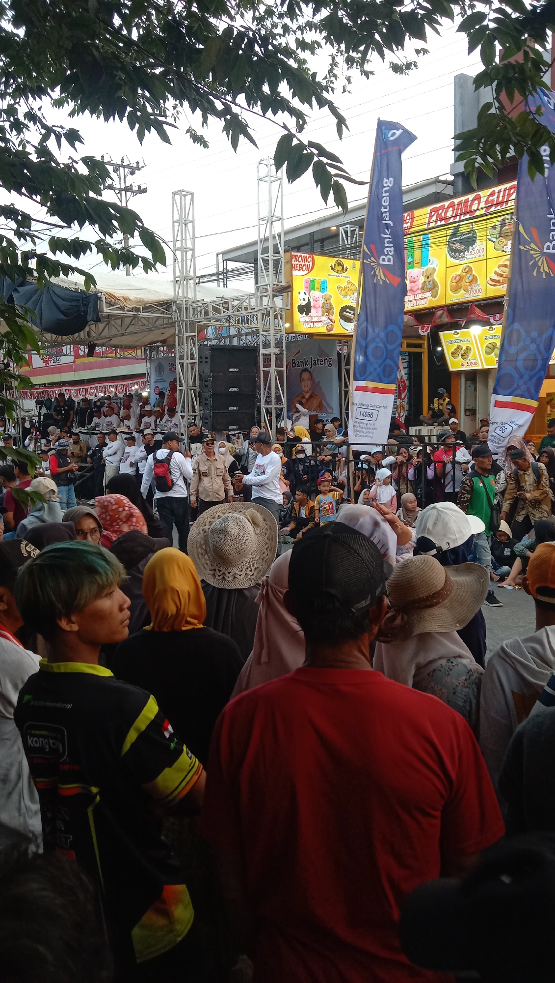 The image captures an outdoor event, likely a festival or market, in Jatisawit, Indonesia, as indicated by the "Bank Jateng" banners which are associated with Bank of Central Java. In the foreground, a crowd of people are gathered, many facing towards a stage area. The people are dressed in casual clothing, with a variety of headwear, including hats and hijabs.
Behind the crowd, a stage structure with scaffolding and speakers is visible. Several banners with promotional messages and logos, including "PROMO HEBOH" and "PROMO SUPER," are displayed on buildings and poles, suggesting commercial activity or sponsorships. A large banner featuring a portrait of a man with the text "Bank Jateng" is prominently displayed. The environment appears to be an urban street or public square, with trees and overhead wires suggesting it's outdoors. The lighting suggests it could be late afternoon or early evening. The overall scene depicts a lively public gathering with a focus on a central event or performance area.