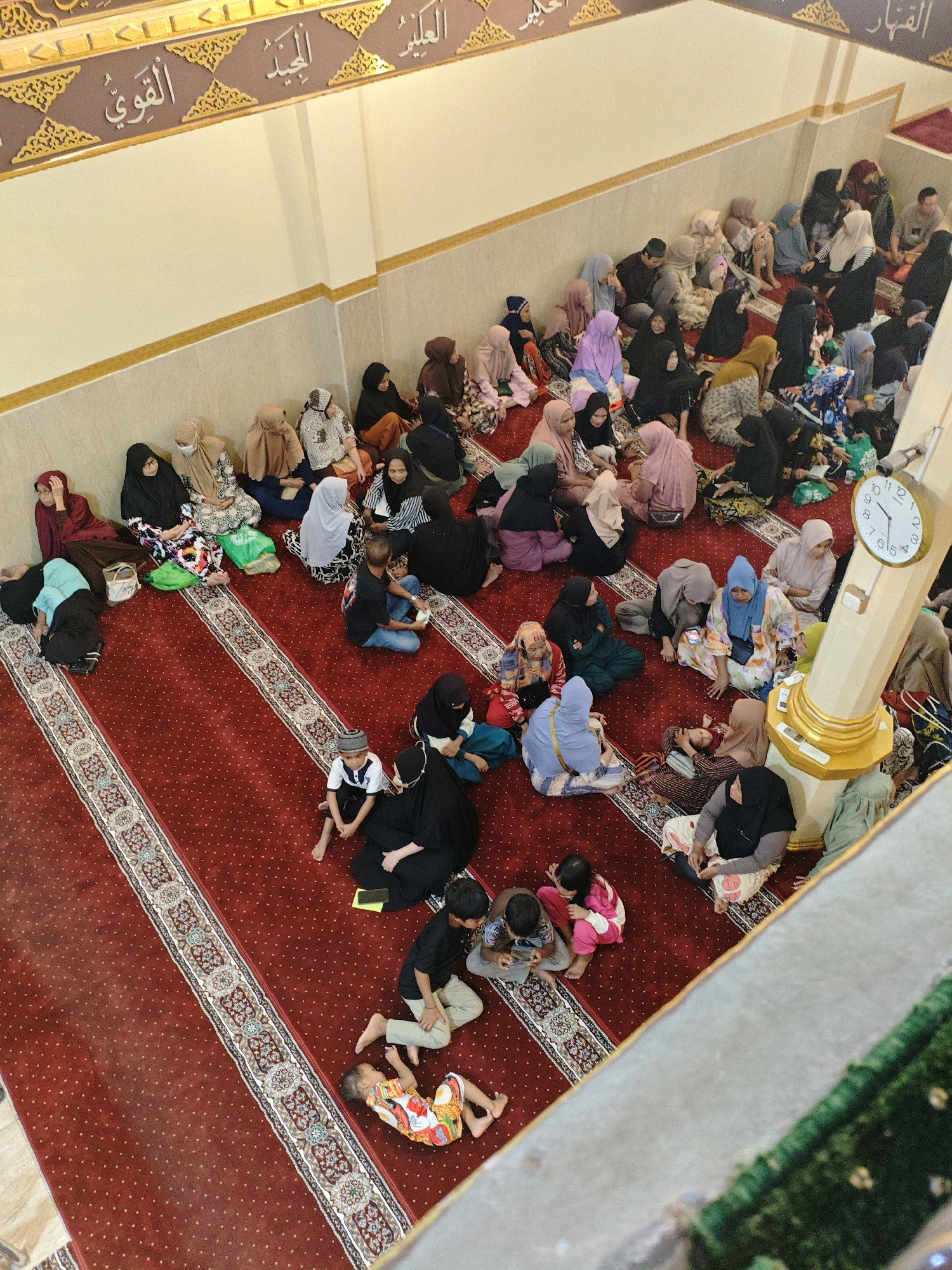 The image shows a crowded indoor scene, likely a mosque, with a large number of people, mostly women and children, sitting on a red carpeted floor. The carpet has a decorative patterned border. Many individuals are wearing headscarves and modest clothing. Some people appear to be in prayer or listening to a speaker, while others are interacting or looking at their phones. A large clock is visible on a pillar in the foreground, indicating the time. The architecture suggests an Islamic place of worship, with Arabic calligraphy on the wall. The overall impression is of a community gathering. The location is identified as Teddaopu, Indonesia.