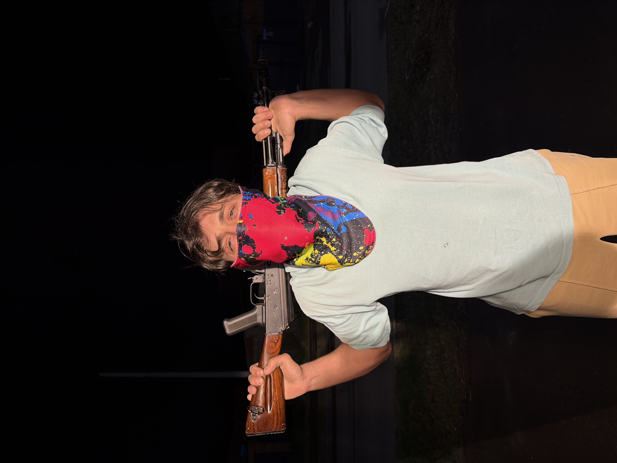 A person wearing a red, black, yellow, and blue patterned bandana partially covering their face is holding an AK-47 assault rifle. The person is wearing a light blue short-sleeved shirt and tan pants. The background is dark, with a hint of greenery suggesting an outdoor setting. There is no indication that this image was taken in Beryozovka, Russia.