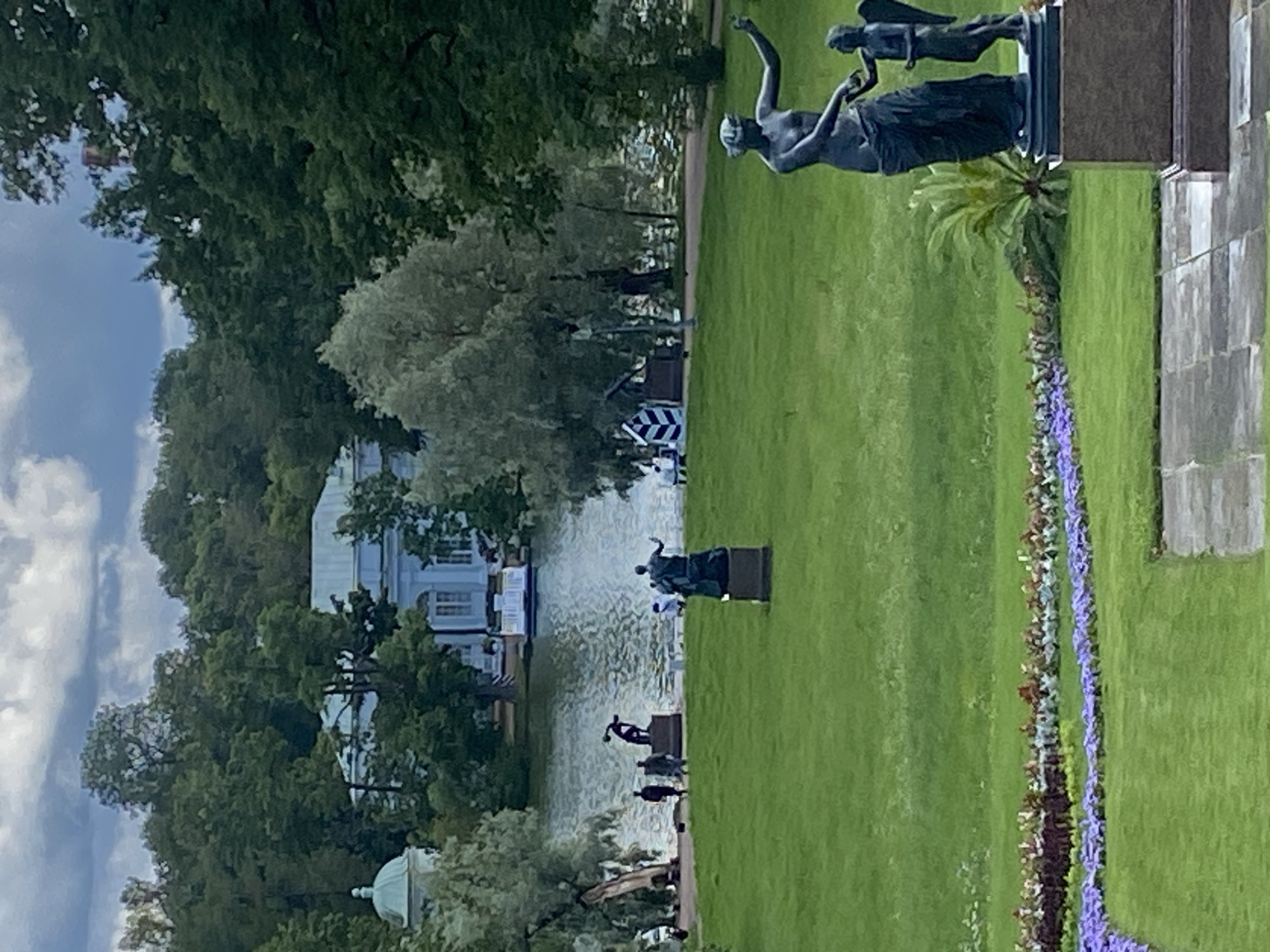 The image depicts a section of the Pavlovsk Park in Pavlovsk, Russia, a suburb of Saint Petersburg. In the foreground, a dark bronze sculpture of a woman holding the hand of a small winged figure stands on a pedestal. To the left of this sculpture, a garden bed with rows of purple and red flowers is visible. The middle ground features a vast expanse of green lawn, dotted with other sculptures. One sculpture, located further back on the left, depicts a muscular male figure in a dynamic pose. Several individuals are walking on the lawn near this sculpture. Beyond the lawn, a body of water, likely a pond or lake, reflects the sky and surrounding trees. On the far side of the water, a light blue neoclassical building with white trim and a colonnade is visible, situated amidst lush green trees. The background is dominated by dense foliage and a cloudy sky. The overall scene suggests a well-maintained park with classical sculptures and architectural elements, characteristic of the Pavlovsk Park.