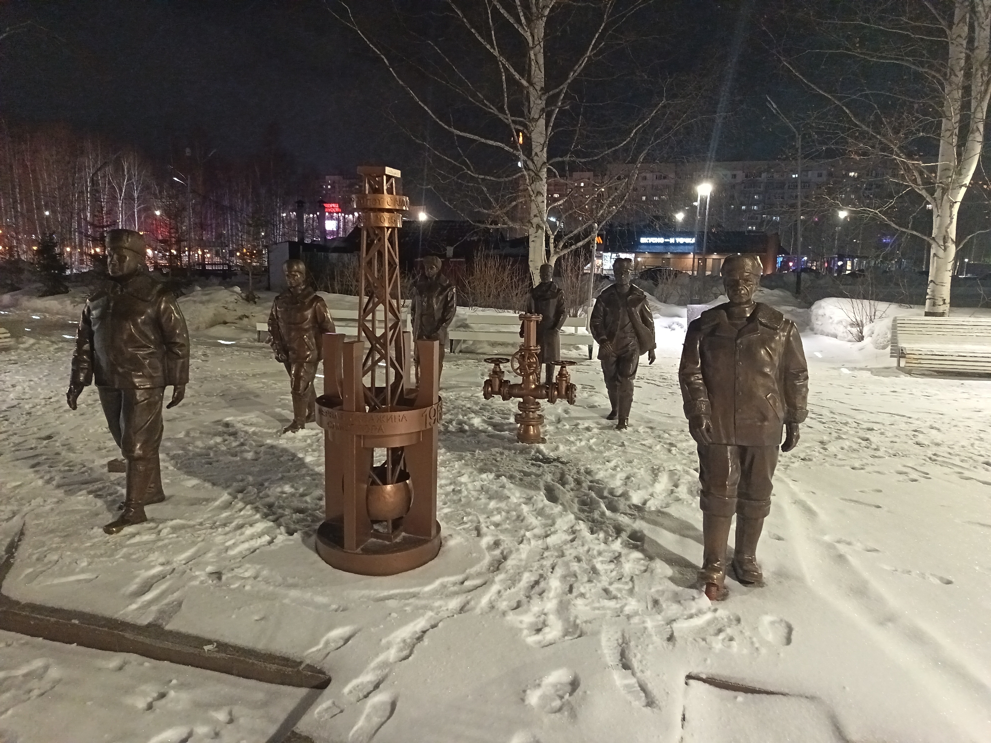 The image depicts a bronze sculpture composition in the city of Nizhnevartovsk, Russia. Six bronze statues of men dressed in thick winter clothing are spread throughout a snow-covered area. Central to the display is a metal framework structure resembling a rudimentary oil derrick or wellhead equipment. The figures appear to be oil workers. In the background, bare trees are seen illuminated by city lights. A multi-story building is visible further back. The overall scene suggests an outdoor commemorative display relating to the oil industry in the Nizhnevartovsk region.
