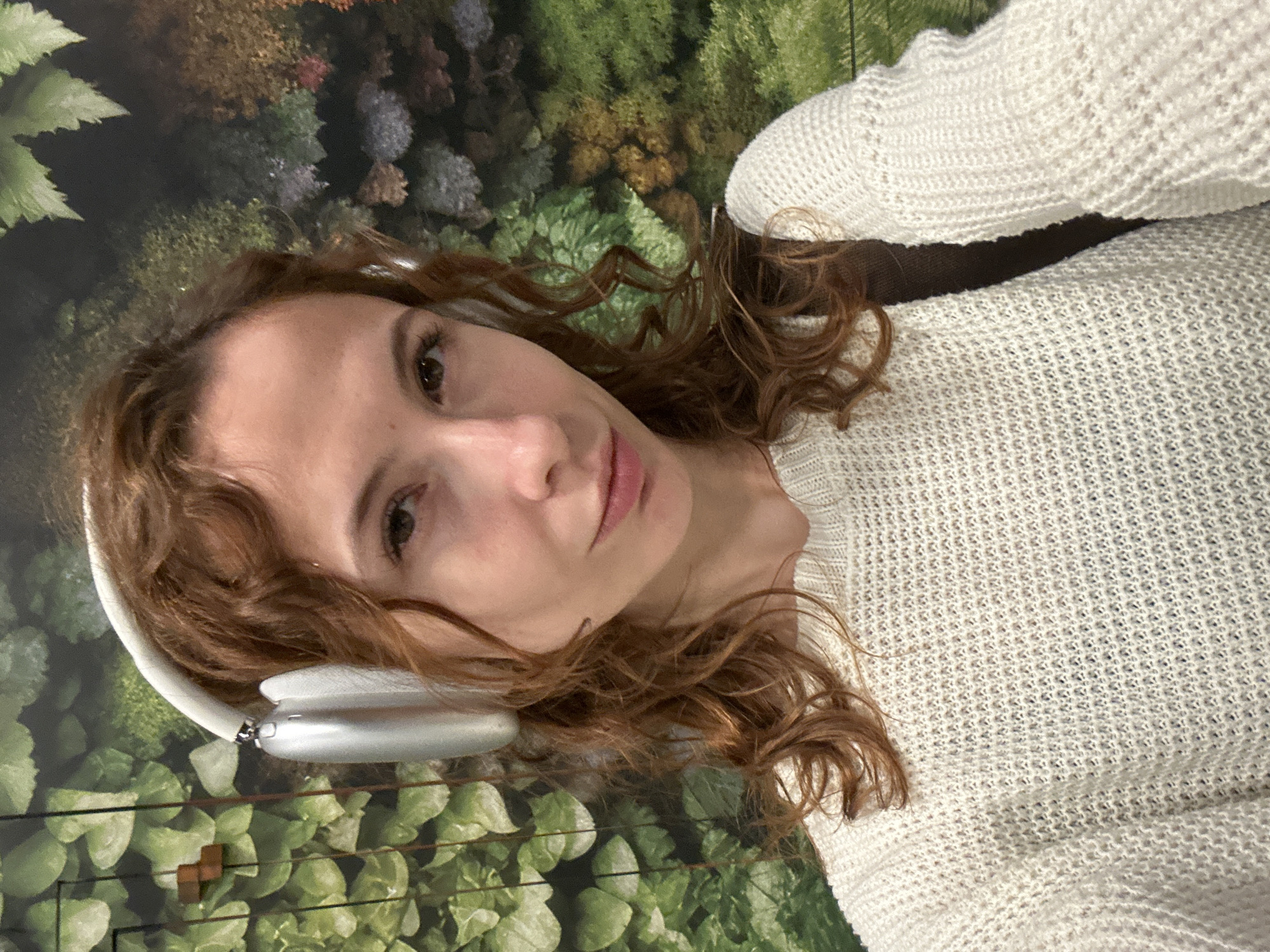 A woman with auburn curly hair and white over-ear headphones is shown in a selfie-style shot, wearing a white knit sweater. The background appears to be a botanical wallpaper featuring a variety of green and brown foliage. Based on metadata, this image was taken inside the "Gatsby" cafe, located at Trg Republike 13, Novi Sad, Serbia.