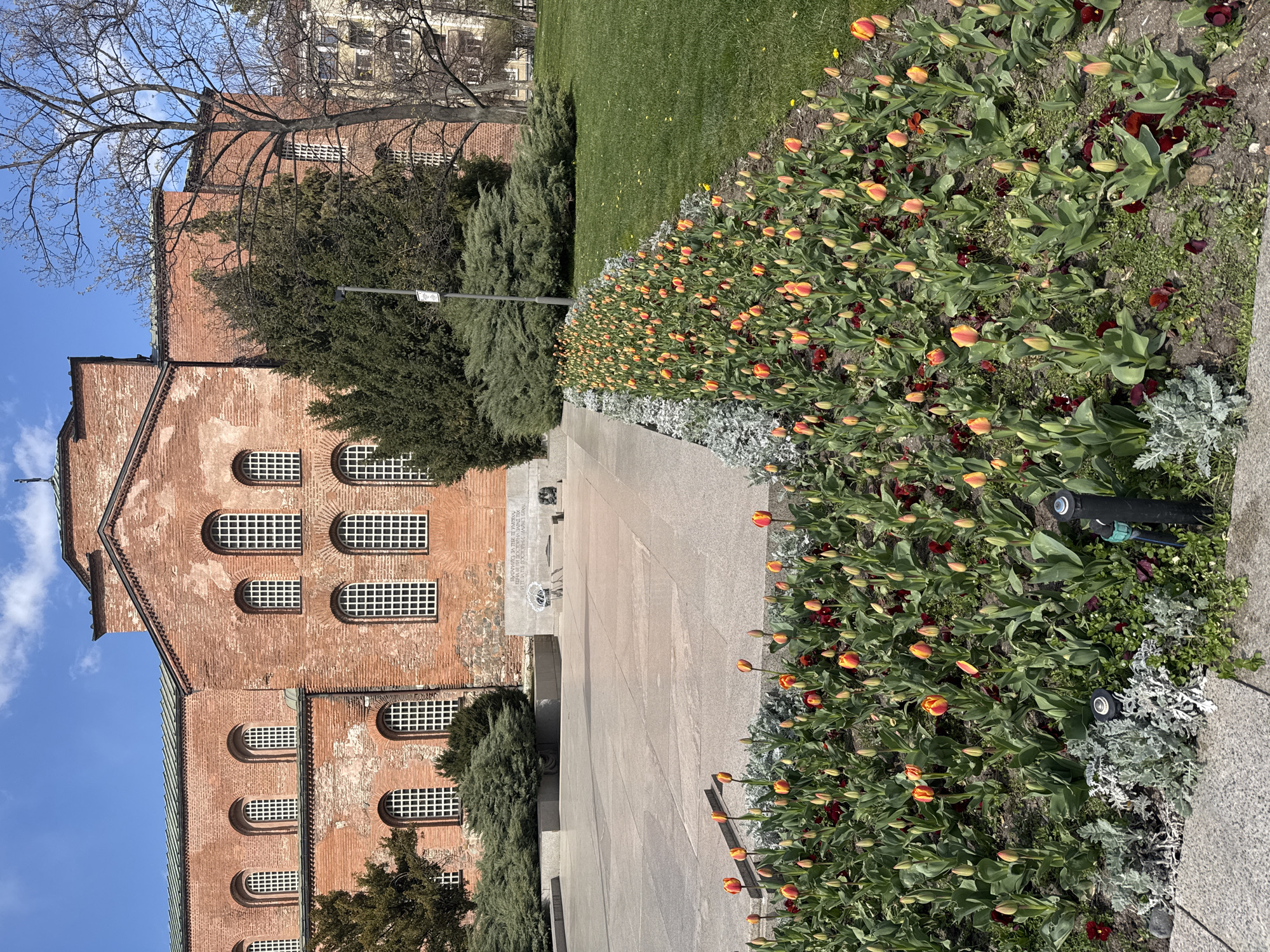 The image captures a section of Sofia, Bulgaria, specifically featuring the Saint Sofia Church and its surrounding grounds. The church, constructed of red brick, stands to the left with visible architectural details such as arched windows. A pathway made of gray stone extends from the foreground towards the church. Adjacent to the pathway, a flowerbed is brimming with colorful tulips and other vegetation. A well-maintained lawn stretches along the top of the frame. A monument sits at the end of the pathway. The sky is blue and clear, with a few clouds visible.