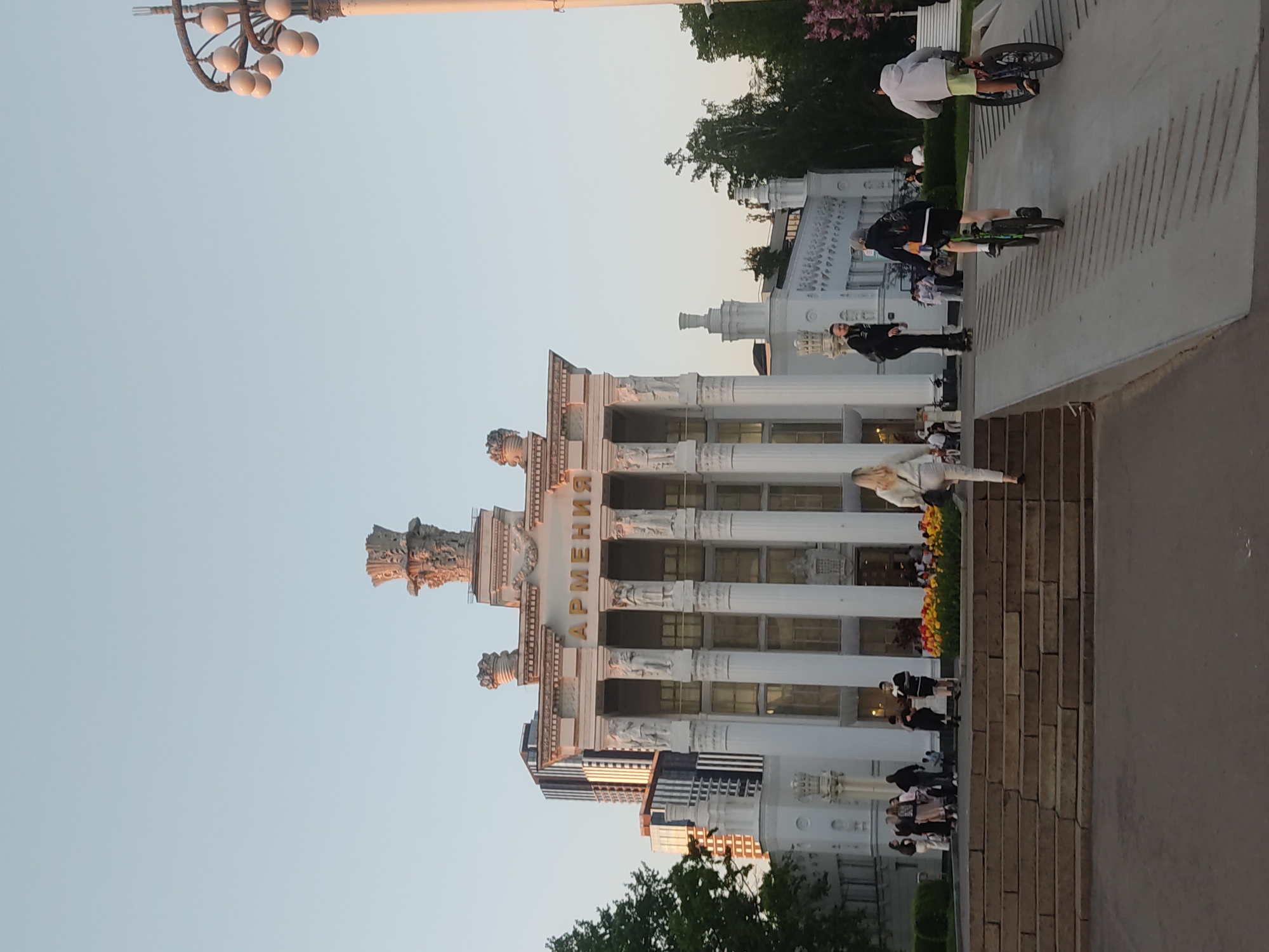 The image shows a building labeled "АРМЕНИЯ" (Armenia) in Moscow, Russia, likely at the VDNKh exhibition park. The building is white with ornate architectural details, including columns and statues. Several people are walking and biking along a paved area next to the building. There are also trees and a lamp post in the background. The sky is clear.