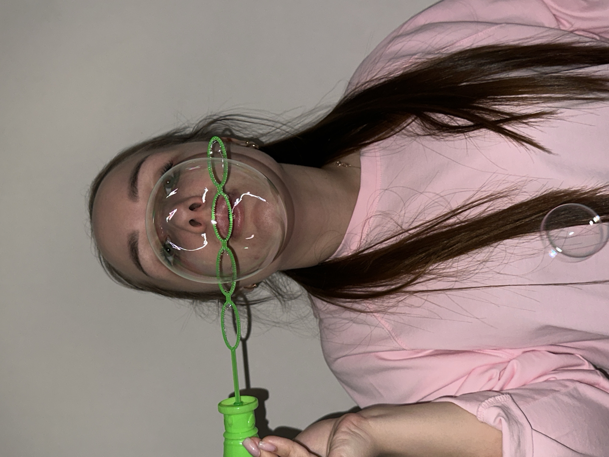 The image displays a young person, likely female, with long, dark brown hair, indoors and engaged in blowing bubbles. Her head is tilted to the left, with her face partially enveloped by a large, transparent bubble she is actively forming. She holds a bright green, plastic bubble wand, featuring a chain-like design of loops, and a matching green bubble mixture container in her right hand. Her mouth is slightly open, focused on the activity, and her eyes are visible through the bubble. She wears a light pink long-sleeved shirt or sweatshirt, and a thin gold necklace is visible around her neck. Her fingernails are painted a light, neutral shade. The background is a plain, light-colored wall, indicating an indoor setting. A smaller, detached bubble floats to the right side of the frame. The overall mood conveys playful concentration. No text is visible in the image. This scene takes place in Chaikovsky, Russia.