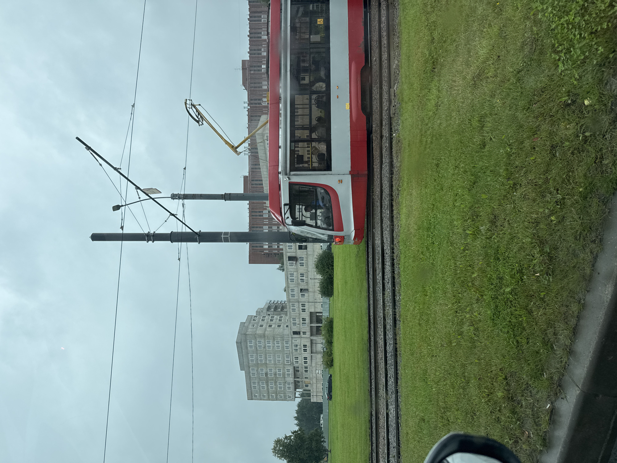 An overcast, daytime, medium shot captures a red and white tram moving left to right on tram tracks, with its pantograph extended to an overhead wire. The tram's windows reveal passengers inside. In the background, a large, multi-story, gray, unfinished or Brutalist style building with many windows dominates the scene. To the right of this building, another, taller, brick building with a red-brown facade is partially visible. Green grass covers the foreground and stretches towards the tram tracks and buildings. Overhead power lines and two tall, dark poles with electrical equipment crisscross the sky. A yellow construction lift with an extended arm is positioned in front of the brick building. A dark sedan is parked on a road or lot near the gray building. The scene appears to be in an urban environment.
