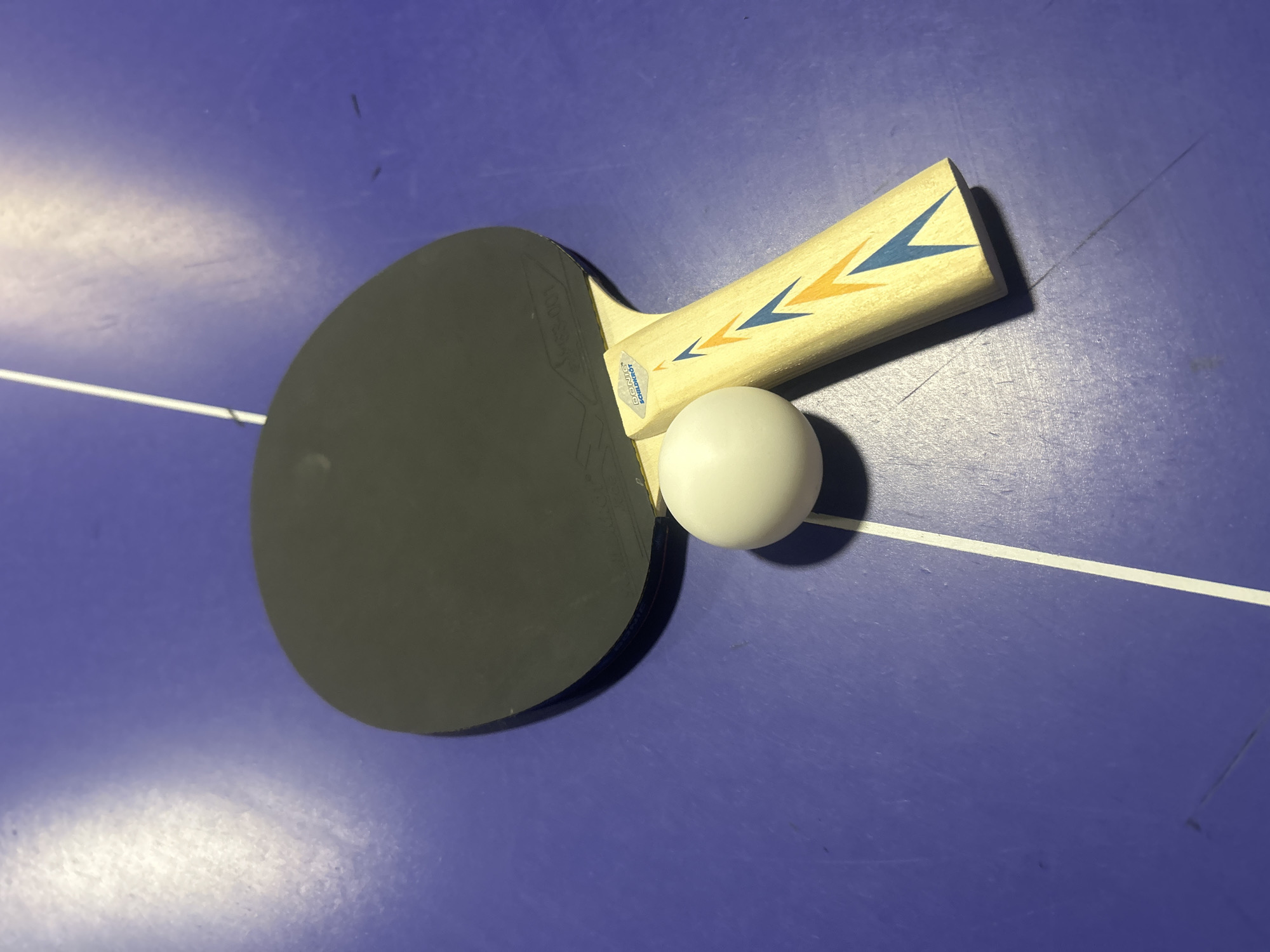 A flat-lay shot depicts a table tennis paddle and a white ball resting on a blue ping pong table, likely in Pleven, Bulgaria. The paddle is positioned diagonally across the frame, with its black rubber surface facing upwards and its wooden handle pointing towards the top right. The handle features a design of blue and orange chevron-like arrows. A small sticker on the handle clearly displays the brand "DONIC SCHILDKRÖT".
The white ping pong ball is situated just below the paddle's handle, touching both the wood and the edge of the rubber. The blue table surface has a distinct white dividing line running horizontally across the middle of the frame. The table appears used, with visible scuffs, scratches, and dust particles scattered across its surface, suggesting a well-played-on area. The lighting, likely artificial, casts soft shadows from the paddle and ball, indicating an indoor setting. No activity is in progress, presenting a static still life of sports equipment.