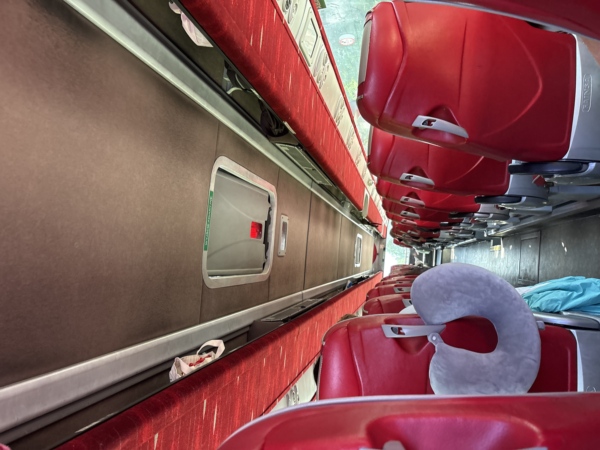 The image shows the interior of a bus, looking forward down the aisle. The seats are red with silver accents, and the overhead compartments are lined with red fabric. There is a neck pillow hanging from a seatback on the left side of the image. The ceiling of the bus has a brown textured surface and a light gray metal trim running along the length of the interior. An emergency exit sign with green lettering and a red light is visible on the ceiling. The scene is forensic and matter-of-fact, depicting the interior of a passenger bus likely in Velyki Pidlisky, Ukraine.