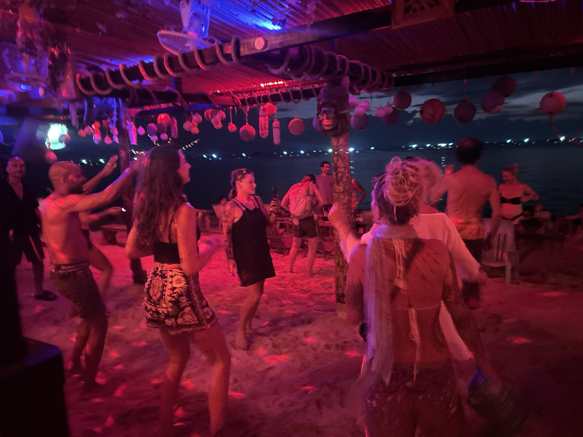 The image depicts a lively night scene at a beachside party in Baan Sri Thanu, Thailand. The environment is illuminated with red and blue lighting, casting a vibrant hue over the sandy ground. A wooden structure overhead is decorated with hanging objects, including circular ornaments and a mask. Several people are present, engaged in various activities such as dancing and standing around. Many appear to be tourists. The backdrop reveals a body of water with lights visible in the distance, suggesting a coastal setting.