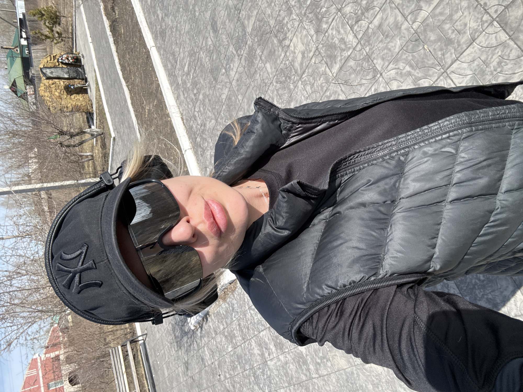 The image captures a person, likely female, in a selfie-style shot or a close-up portrait taken outdoors on a sunny day. The primary subject is positioned in the foreground, facing the camera at a slight upward angle. She wears large, reflective rectangular sunglasses that obscure her eyes, and her lips are visible, appearing full and natural. A black baseball cap, featuring a prominent, stylized black embroidered logo (resembling the "NY" typically associated with the New York Yankees, but highly stylized), covers her head. Her light-colored hair is visible pulled back behind her.
She is dressed in a black ensemble, consisting of a puffy, quilted black vest worn over a long-sleeved black top. A delicate necklace is visible around her neck.
The background reveals an outdoor setting, suggesting a park or memorial area in Borzya, Russia. A patterned paved pathway or sidewalk runs diagonally through the scene. Beyond the path, there's an area with bare trees, indicating late winter or early spring. In the mid-ground, a military-themed monument or memorial stands, covered in what appears to be camouflage netting or textured material, with a funeral wreath placed on top. A small, greenish building is partially visible further in the background to the left. The bright sunlight creates clear shadows, suggesting a midday or afternoon shot.