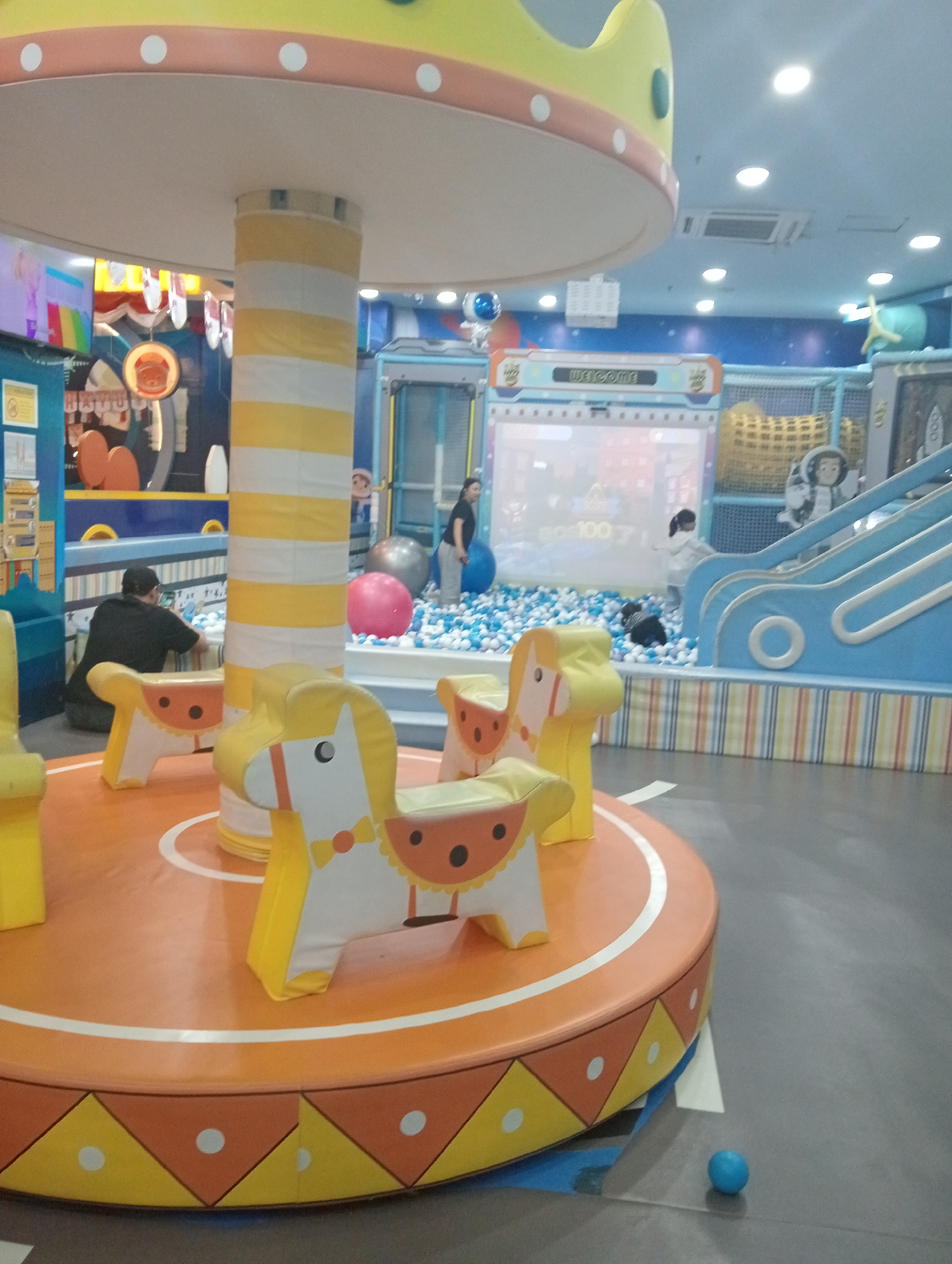 The image depicts an indoor children's play area, possibly a carousel, in Sempusari, Indonesia. In the foreground, a circular platform with a decorative pattern of yellow and orange triangles and white circles is visible. On this platform, there are several stylized white and yellow rocking horses with orange circular accents. A thick, striped yellow and white pole supports a large, yellow, circular structure above.
In the mid-ground, a man wearing a black shirt and a dark baseball cap is seated, facing away from the camera, near the striped pole. To his right, a bright pink inflatable ball rests on the floor. Behind the man and the ball is a large ball pit filled with white and blue balls. A woman in a dark top and light-colored pants stands in the ball pit, and a child in a white outfit is also present near the pit. A blue slide with white railings is visible to the right of the ball pit, with a decorative astronaut figure on it.
The background shows various play structures and decorations, including a large screen displaying colorful graphics and the word "Welcome." The overall environment appears to be a bright and colorful indoor amusement facility. The flooring is a dark gray material. A small blue ball is visible on the floor in the lower right corner of the image.