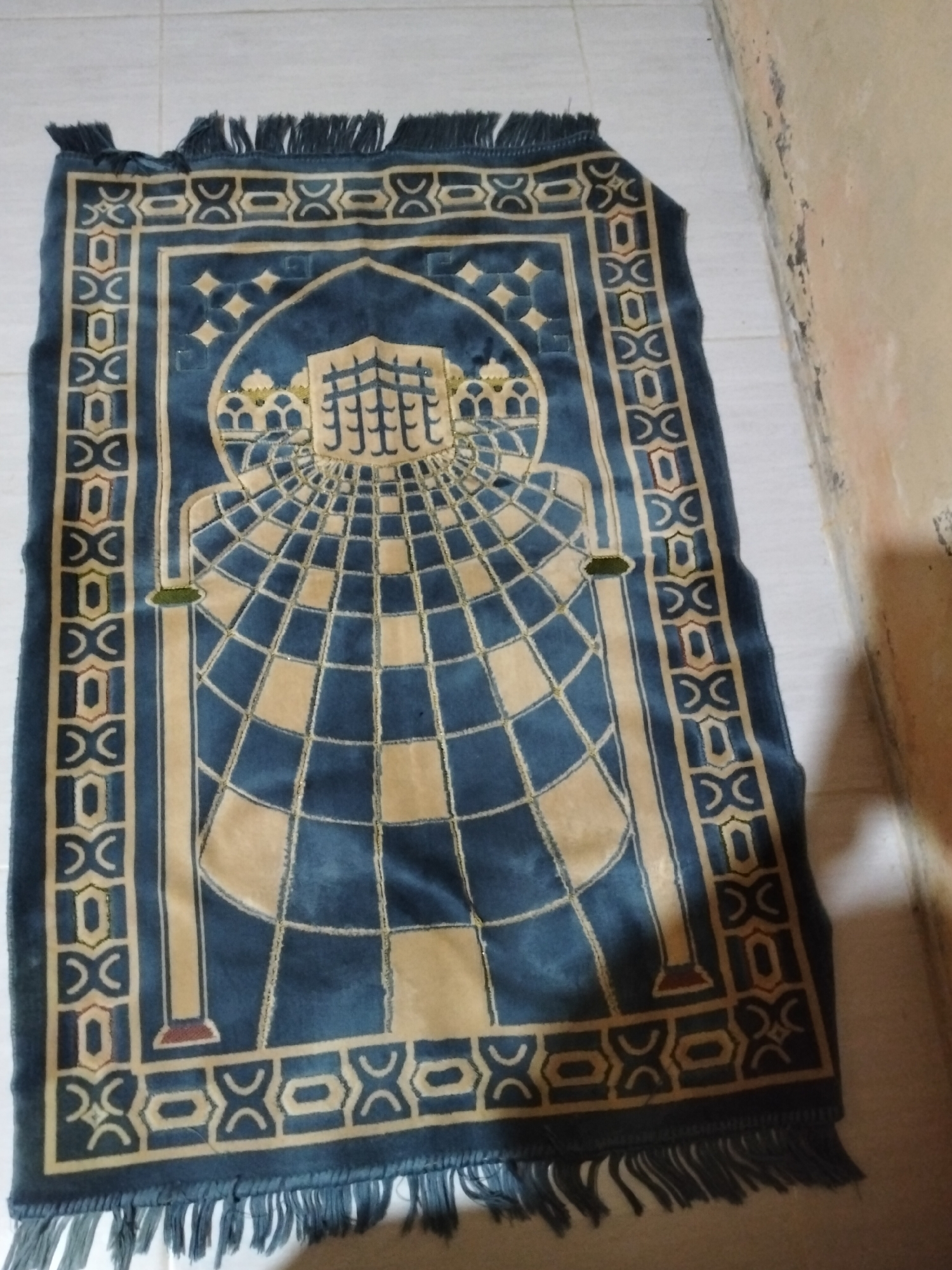 The media file shows a prayer rug lying on a tiled floor. The rug is rectangular with a fringe on both ends. It features a central design of a checkered pattern that converges towards a depiction of the Kaaba in Mecca at the top. The rug is primarily blue and beige, with intricate geometric patterns along its borders. The setting appears to be indoors, possibly a home, given the tiled flooring and a wall visible on the right side of the image. There is no visible text in the image, nor any people or indication of an ongoing activity or event. The lighting suggests it might be daytime. There are no discernible weather cues or strong emotional indicators. The location context provided is Payagaboh, Indonesia.