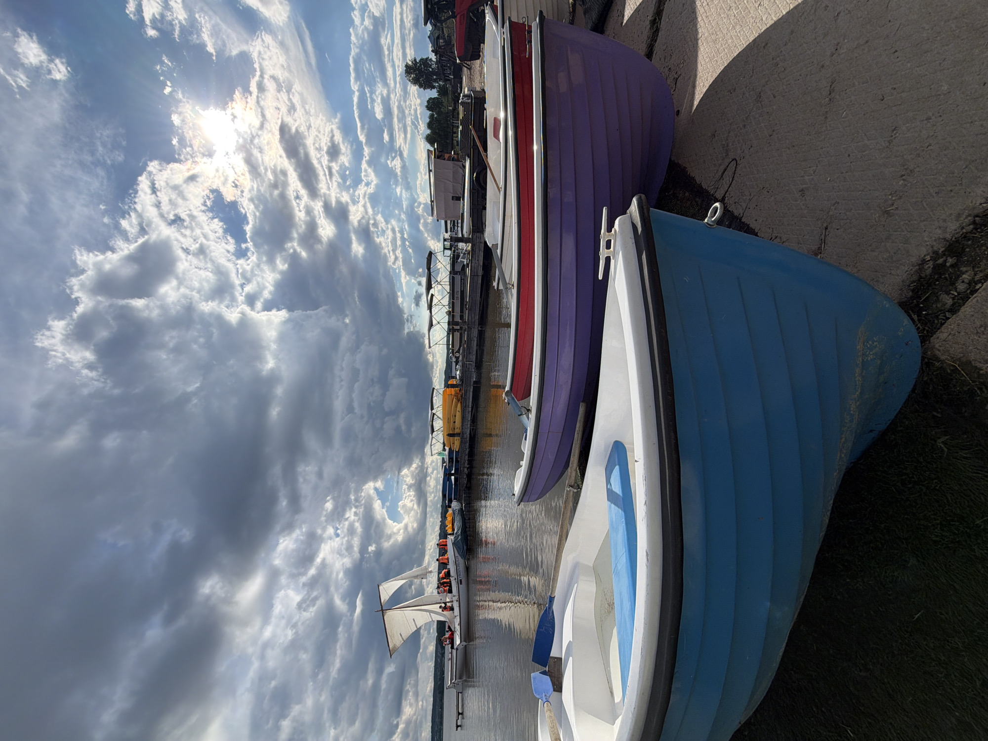 The image depicts a scene at a dock, possibly in Votkinsk, Russia, given the prompt. Several small boats, painted in blue, purple, and red, are pulled up onto a concrete landing. Beyond the boats, a body of water extends to the horizon. A sailboat with passengers wearing orange life jackets is visible on the water. In the background, there are docks, buildings, and trees. The sky is filled with numerous clouds, with the sun shining through, creating a bright, diffused light across the scene. The photo is taken from a low angle, emphasizing the boats in the foreground and creating a sense of depth leading to the horizon.