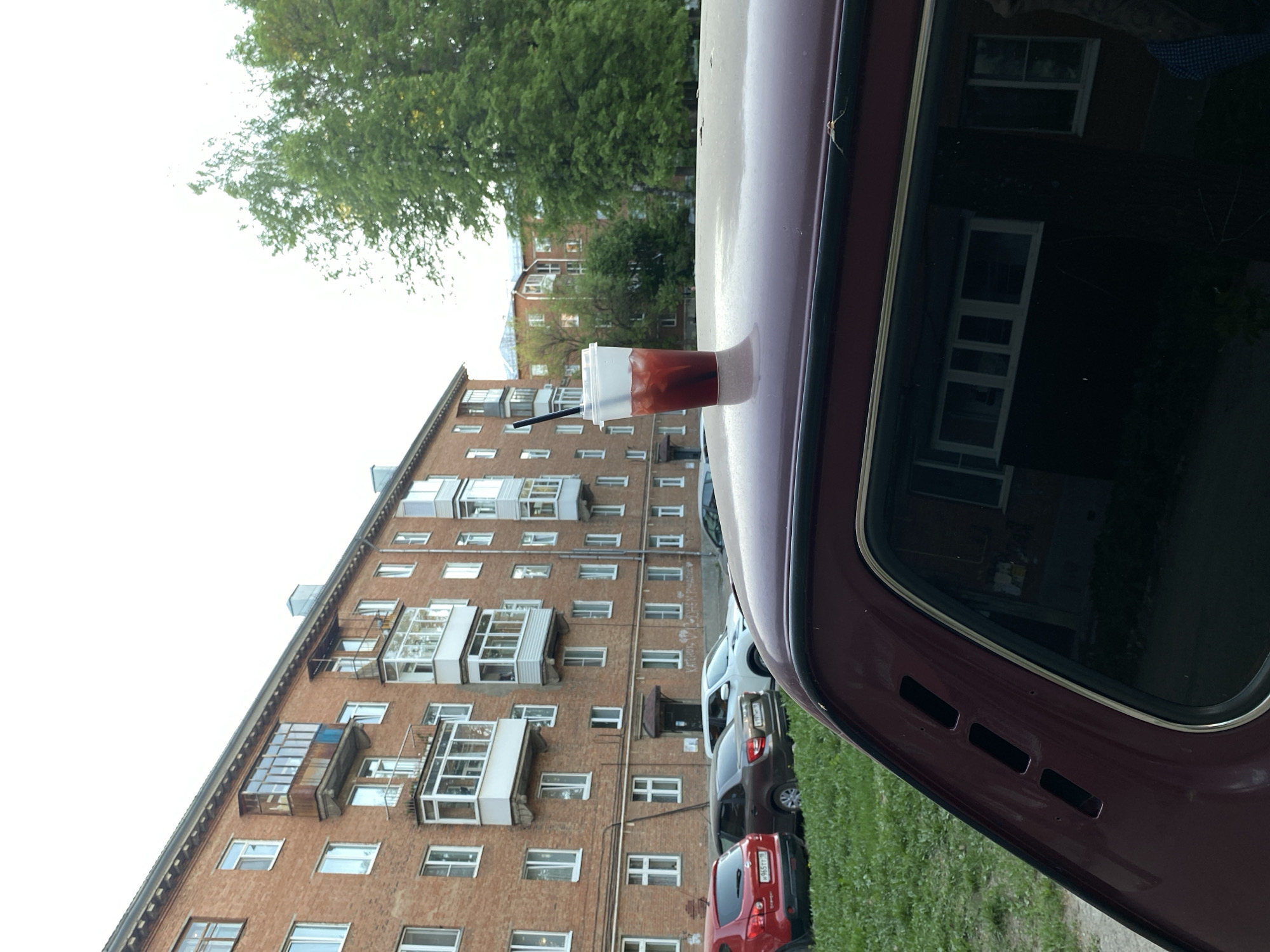 Here's a description of the image:
The image shows a red drink in a clear plastic cup with a black straw, perched on the roof of a maroon car. The open car door is visible, showing reflections. In the background is an apartment building with multiple windows and balconies, and several cars parked in front. A green tree is also visible. The photo appears to be taken in the city of Votkinsk, Russia, based on the architecture of the building and parked cars, as well as the style of the balconies. The building seems to be an older, multi-story residential building.