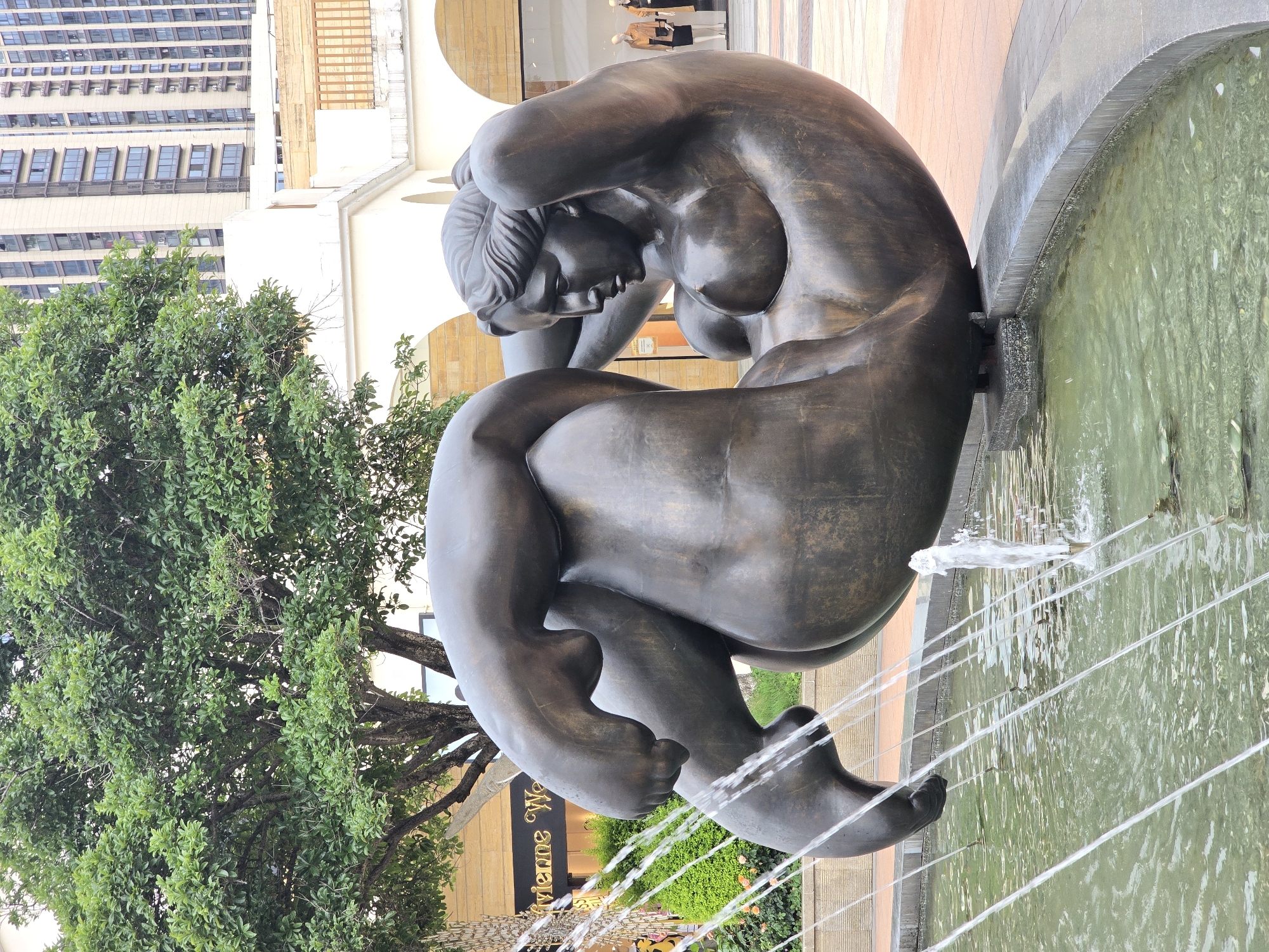 This image depicts a bronze sculpture of a reclining nude woman situated in a public fountain area. The sculpture is centrally located in the frame, with its bronze surface showing a patina of age. The woman is posed in a curled position, with her head resting on her hands, which are behind her head. Her body is curved, and her legs are drawn up. Water sprays from multiple jets around the sculpture, creating a dynamic effect in the shallow pool.
In the background, there is lush green foliage from a large tree on the left side of the frame, and a modern building with many windows on the right. Part of a store sign, "Vivienne West..." is visible on the left, suggesting a commercial area. Mannequins in a storefront can be seen in the far right background. The ground surrounding the fountain is paved with reddish-orange tiles.
This scene is located in Haining, China, specifically at the Haining Longfor Paradise Walk.