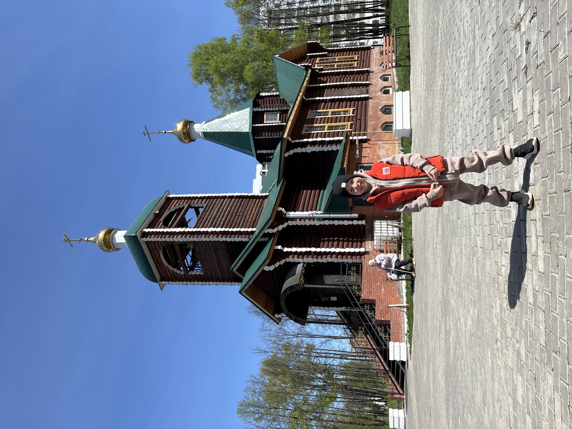 A person stands on a paved surface in front of a wooden church on a sunny day. The church has green roofs and golden domes with crosses. The pavement is light gray and has a brick-like pattern. Trees are visible in the background, and the sky is clear blue. An elderly person sits on a bench near the building. The person in the foreground wears a red vest and a dark cap. The location is likely in the city of Chaikovsky, Russia.