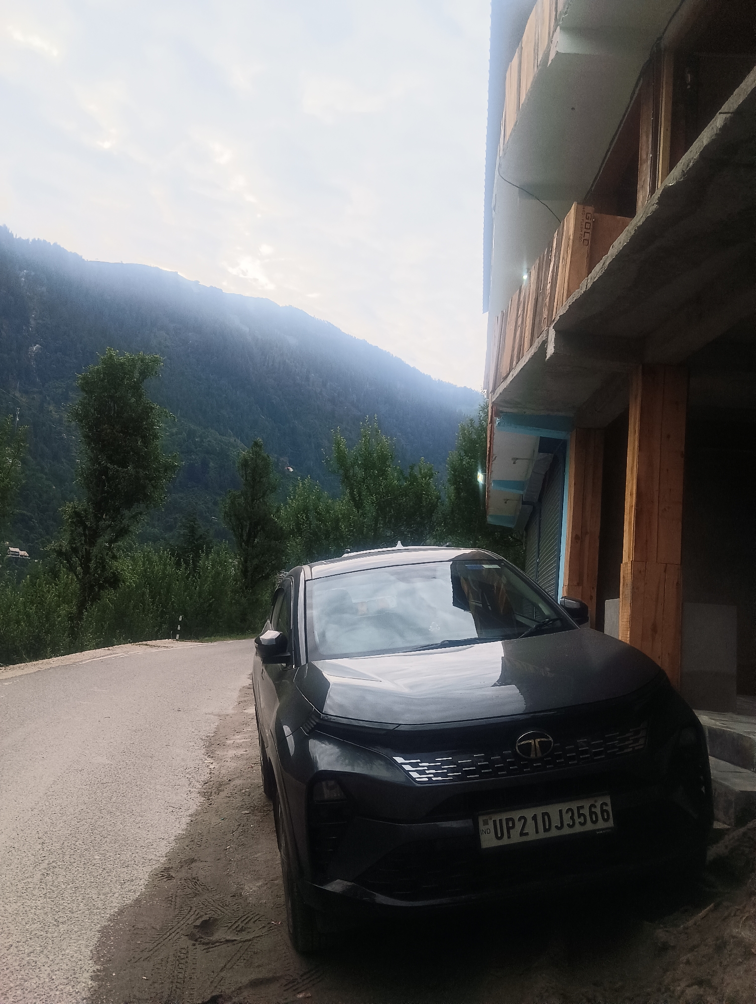 The image depicts a dark-colored SUV parked on a dirt and asphalt road in the vicinity of a multi-story building under construction. The car's license plate reads "UP21DJ3566", indicating it is registered in Uttar Pradesh, India. The background features a lush, green mountain landscape with trees and a hazy sky. The building appears to be made of wood and concrete, with unfinished elements. The road is wet in places, suggesting recent rain. The overall scene suggests a rural or semi-rural setting, possibly in the Himalayan region of India.