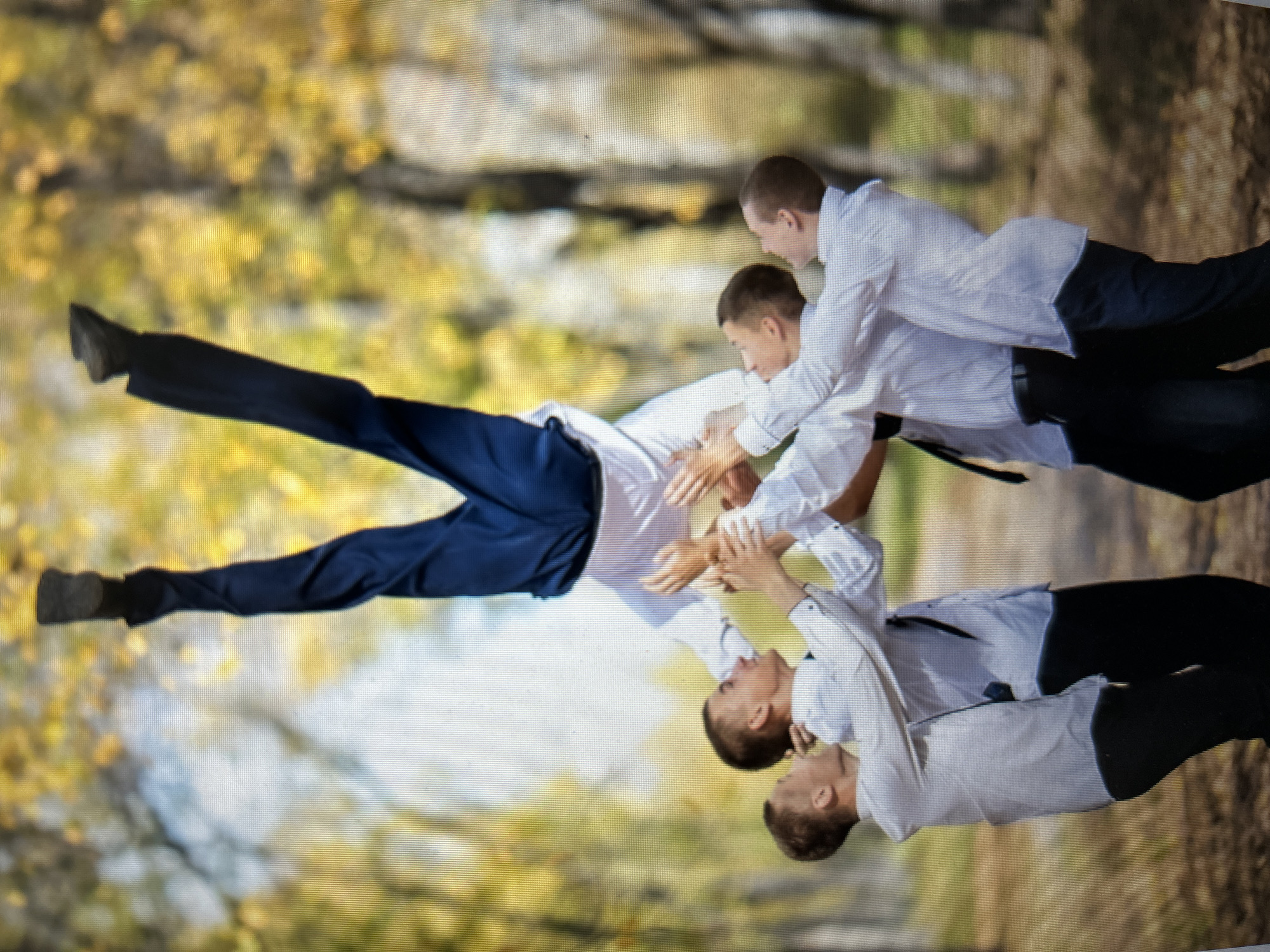In an outdoor park setting, likely autumn given the yellow foliage, five young men in white button-down shirts and dark trousers are participating in an acrobatic stunt. One man is being supported upside down by the other four, with his legs extended high in the air. The man in the air is wearing dark blue pants. The other four men are positioned around him, with their hands clasped to support his body. The background is blurred, showing trees with yellow leaves and a path on the ground. This scene is unlikely to be located in the city of Kiselyovsk, Russia, as there are no identifiable landmarks or context to suggest this specific location.