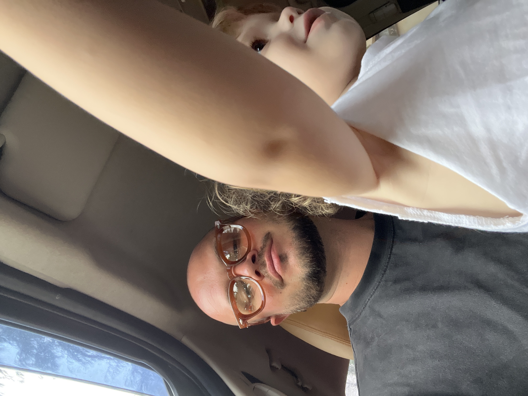 A bald man wearing glasses and a black t-shirt is in the driver's seat of a car, looking at the camera. To his right, a child with blond hair, wearing a white shirt, is visible. The child's arm is in the foreground, partially obscuring the man's face. The car's sunroof is open, letting in natural light. The interior of the car is visible, with the dashboard and a tan leather seat. The background is blurred and indistinct.