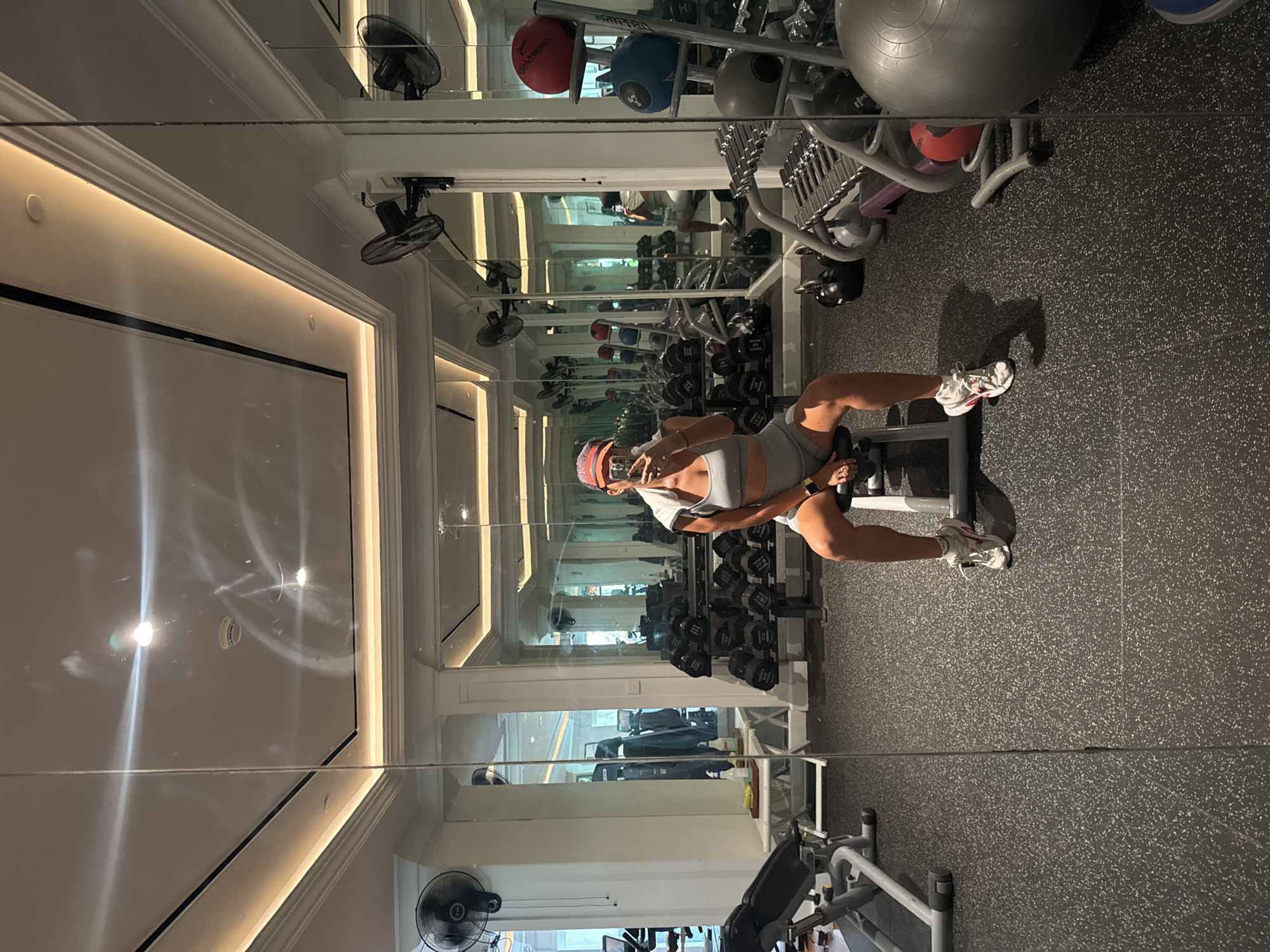 A person is using a weights bench to do seated bicep curls in a gym mirrored on one side. The gym is filled with equipment including dumbbells, kettlebells and medicine balls. There are ceiling fans and air conditioning visible in the mirrors. The location is Kuta Selatan, Indonesia.