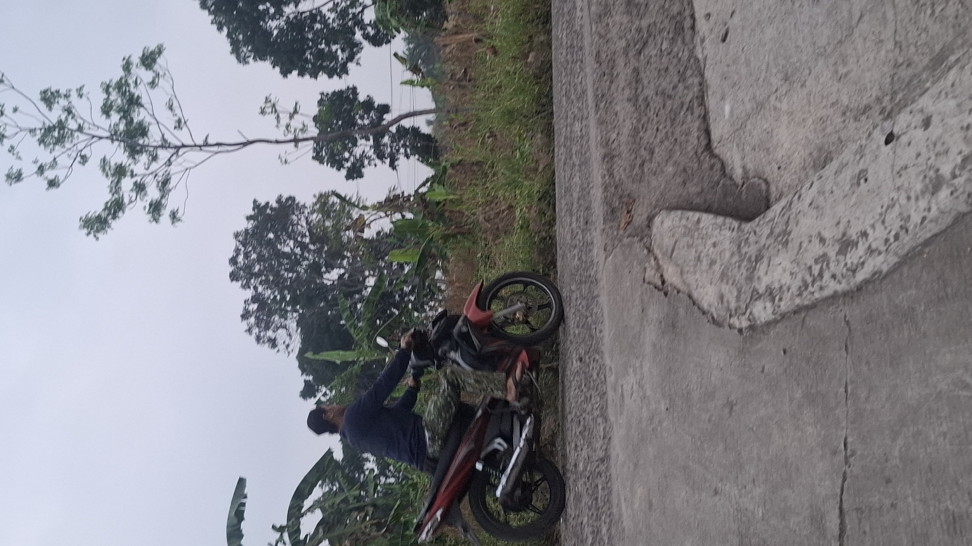 A person is riding a red and black motorcycle on a paved road in Cibinong Hilir, Indonesia. The rider, wearing a dark long-sleeved shirt and camouflage pants, is positioned on the left side of the motorcycle. The motorcycle is facing left and appears to be in motion. The road surface is concrete with some visible cracks and a raised concrete edge. To the right of the road is a grassy embankment with various types of vegetation, including tall trees and banana plants. The sky is overcast.