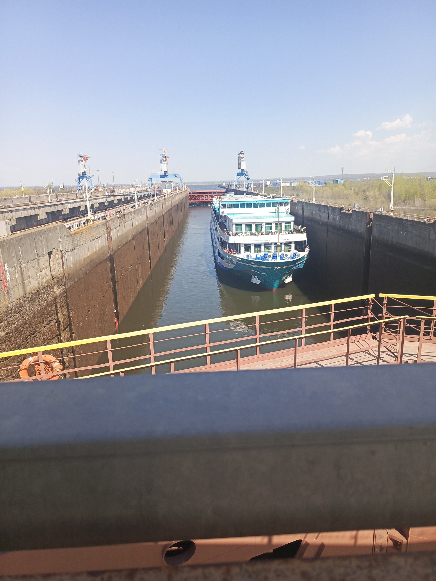A teal and white passenger riverboat is positioned within a concrete lock, enclosed by tall, vertical walls. The lock is part of a waterway in Chaikovsky, Russia. The waterway is edged by cranes and metal walkways. The sky is clear with sparse clouds. An orange life preserver is visible on the railing. The image is taken from an elevated perspective behind a grey metal railing.