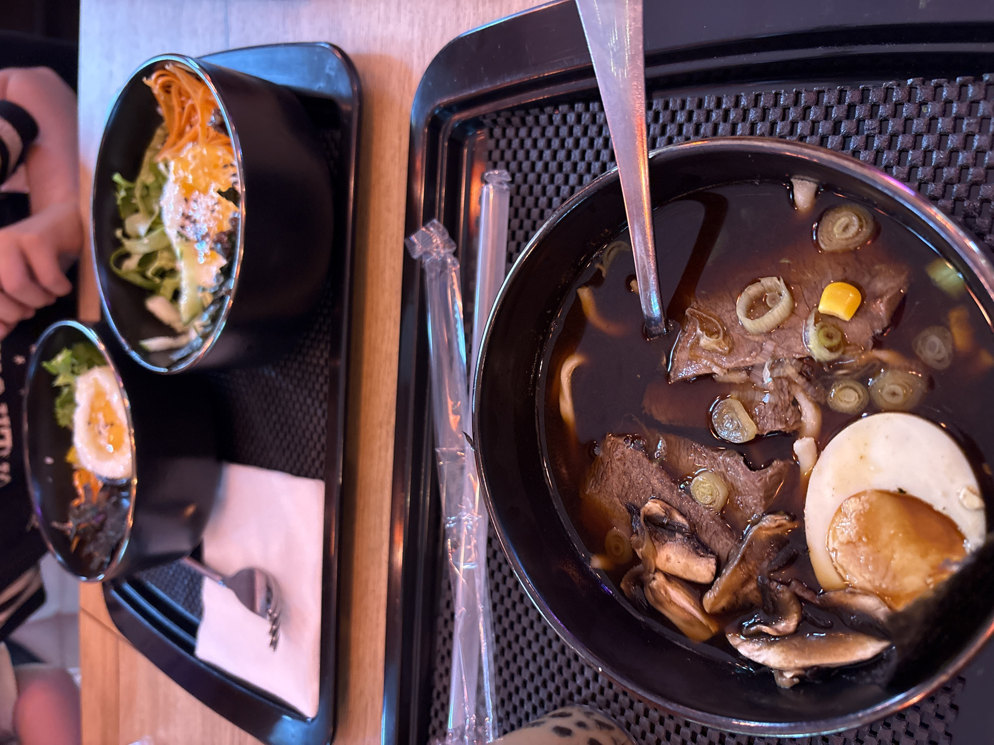 The image shows two black bowls on a black tray. One bowl, in the foreground, contains a dark broth with noodles, sliced beef, mushrooms, a hard-boiled egg, and green onions. A silver spoon rests on the rim of this bowl. Behind this, on another black tray, are two more bowls. The bowl on the left contains a colorful dish with rice, seaweed, and a fried egg. The bowl on the right contains a similar dish with rice, vegetables like shredded carrots and greens, and a fried egg. There are two plastic-wrapped straws on the tray in the foreground. The setting appears to be a restaurant with a wooden table visible in the background. The overall impression is that of a meal being served. The location in Saint Petersburg, Russia, is not discernible from the image.