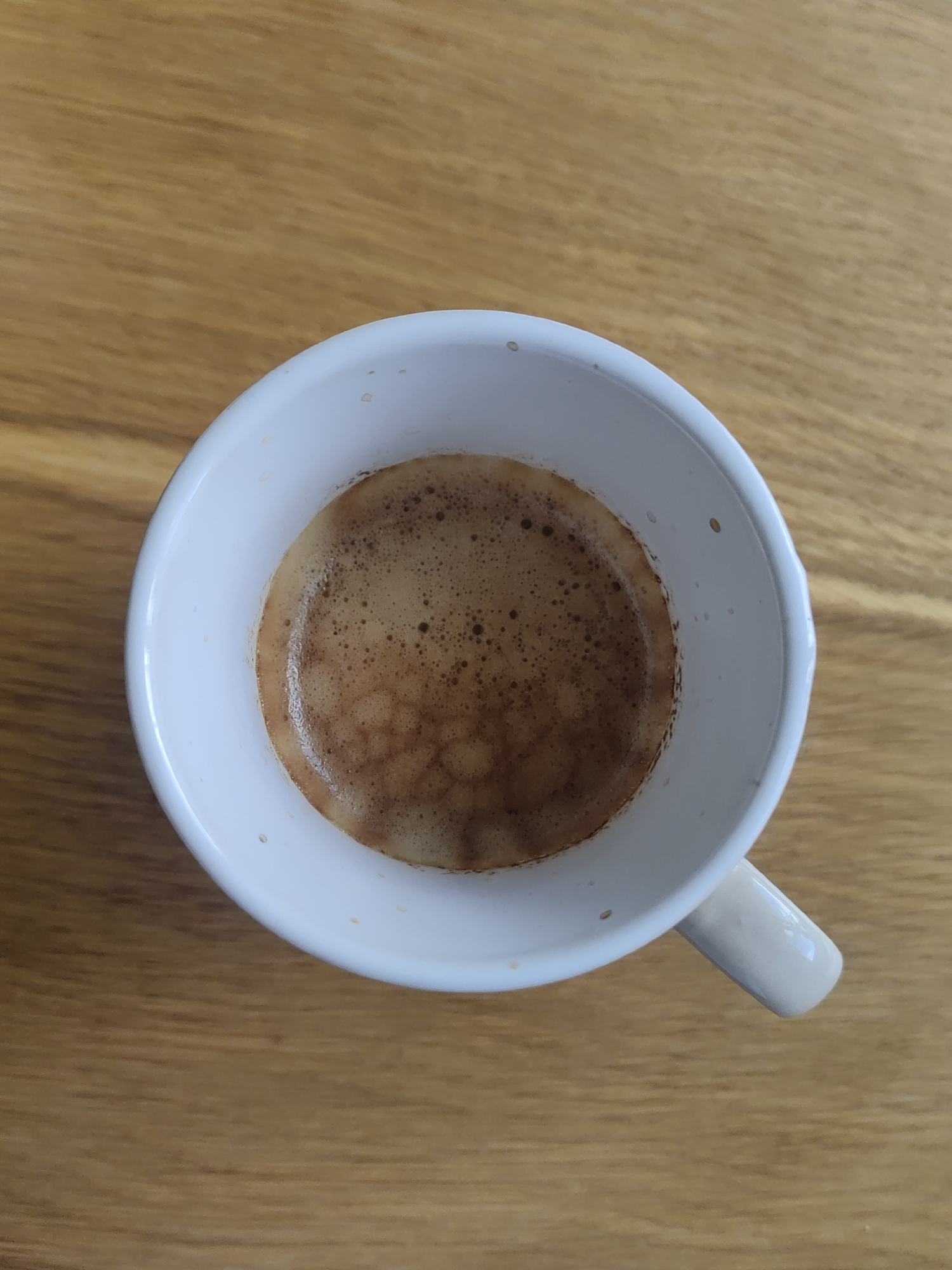 The image shows a close-up, top-down view of a white ceramic mug filled with dark espresso coffee. The coffee has a thick, foamy crema with a pattern of lighter and darker brown tones. The mug is placed on a wooden surface with a visible grain pattern. The setting appears to be indoors, on a table. There are no people or text visible in the image. The lighting suggests it could be daytime.