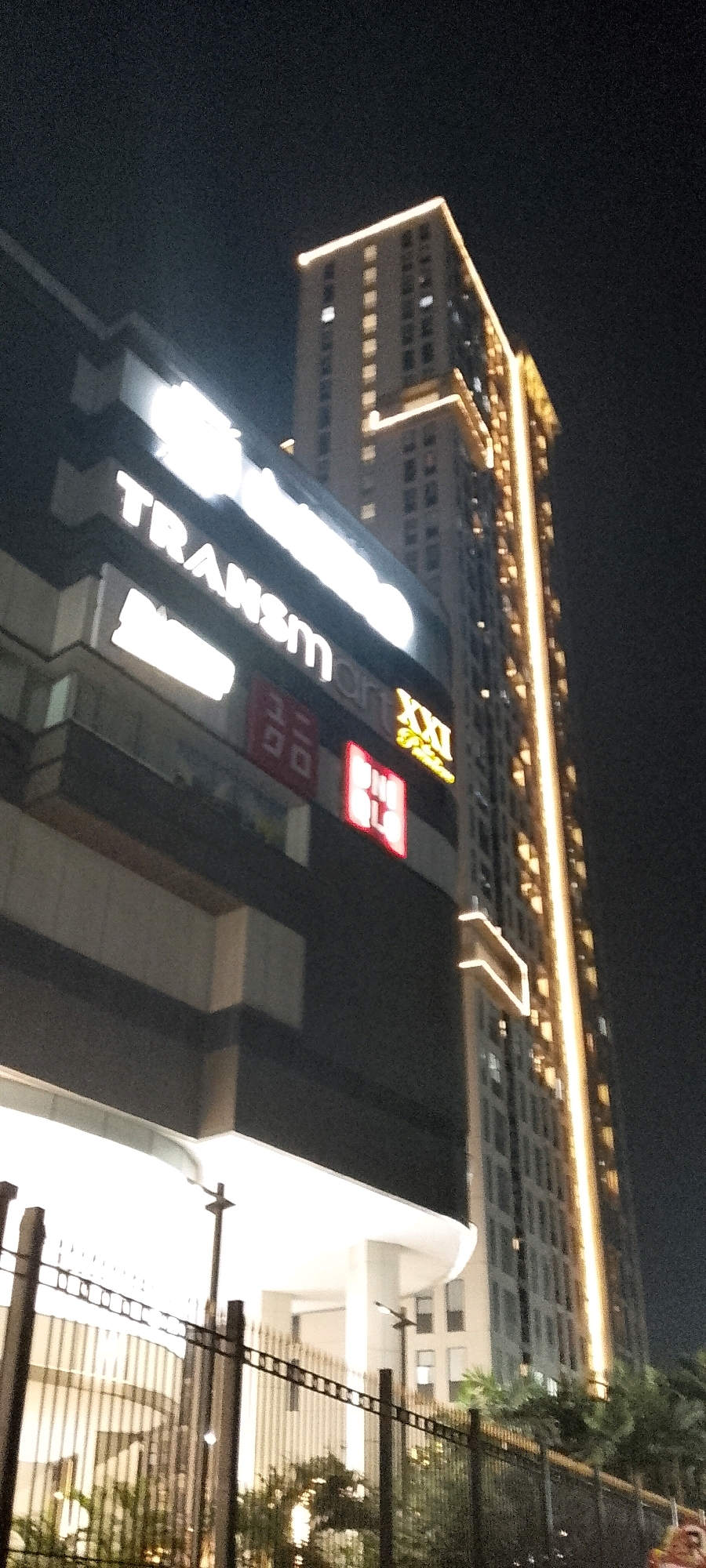 The image shows a night view of a modern building complex in Depok, Indonesia. A tall, illuminated skyscraper stands prominently in the background, its windows lit up, and vertical lighting accentuating its structure. In the foreground, a multi-story building displays large, illuminated signs, including "Transmart" and "XXI," indicating a shopping mall or entertainment center. The lower part of the foreground building features a brightly lit white facade, possibly an entrance or shopping area. A black metal fence runs across the bottom of the frame, partially obscuring a view of the illuminated building entrance and some greenery. The scene is dark, typical of a night setting, with the artificial lights of the buildings being the primary light sources.